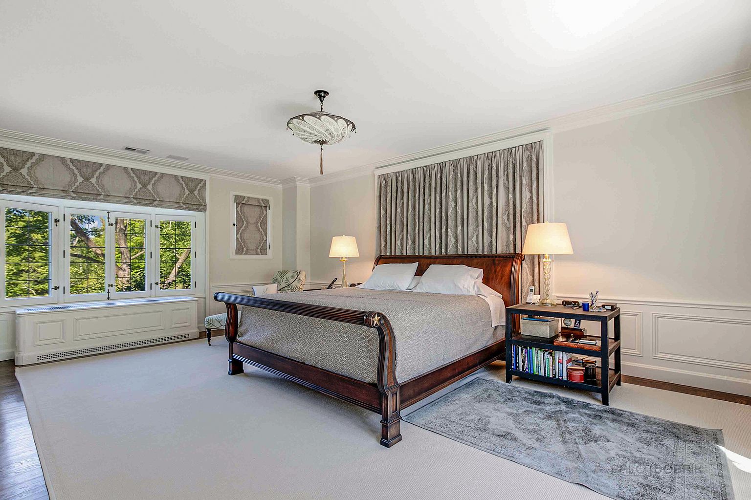 This is a well-lit primary bedroom featuring a large wooden sleigh bed with a patterned bedspread and white pillows. The room has a neutral color palette with white walls and carpeting, complemented by patterned window treatments and a decorative ceiling light fixture. A dark-colored nightstand with books and a lamp sits beside the bed, adding a touch of sophistication.