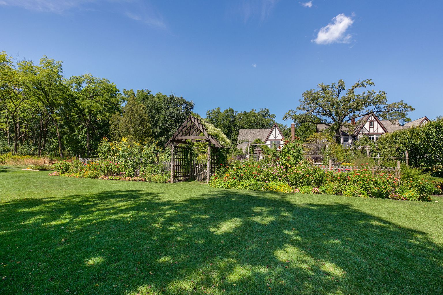 This image showcases a beautifully landscaped yard and garden, featuring a wooden pergola entrance, vibrant flower beds, and a well-maintained lawn. In the background, Tudor-style houses add to the charm and character of the property. The scene is bathed in sunlight, creating a welcoming and picturesque outdoor space.
