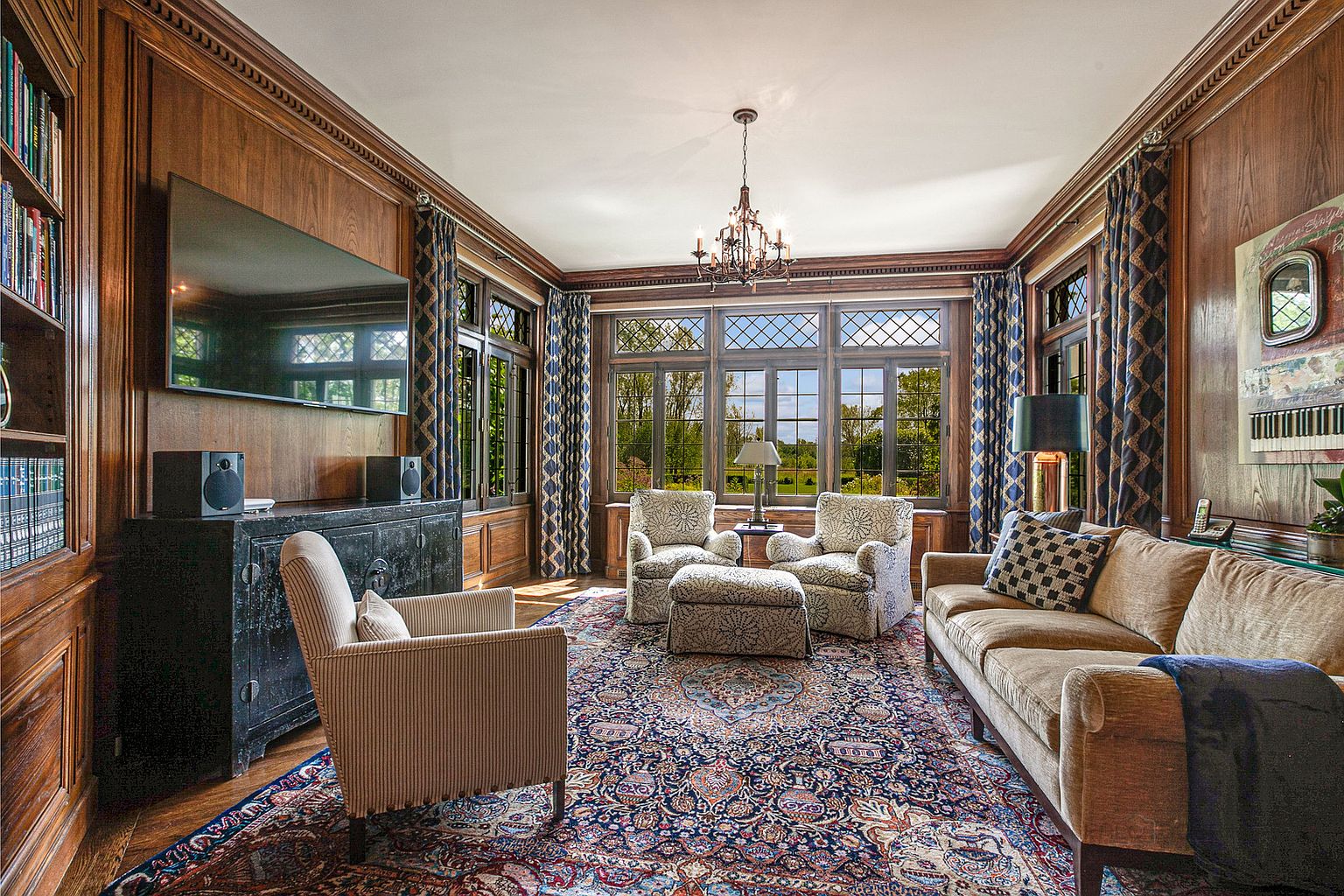 This is a luxurious living room featuring rich wood paneling, a large area rug, and comfortable seating arrangements including armchairs and a sofa. The room is well-lit by a chandelier and natural light streaming through large windows, which offer a view of the outdoors. A large television is mounted above a dark cabinet, flanked by speakers.