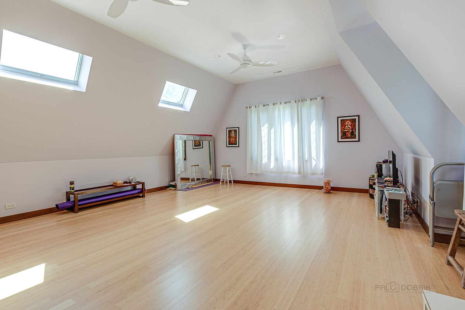 This is an interior shot of a home gym featuring light wood flooring and light purple walls. The room has two skylights and a ceiling fan, providing ample natural light and ventilation. The space is equipped with a mirror, a small bench, and a yoga mat, suggesting a focus on fitness and wellness.
