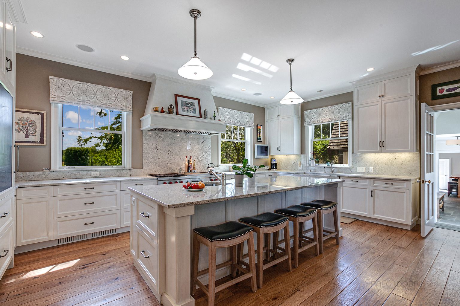 This is a bright and spacious kitchen featuring white cabinetry, granite countertops, and stainless steel appliances. A large central island with seating provides ample workspace and a gathering spot. Natural light floods the room through multiple windows, highlighting the hardwood flooring and creating a warm, inviting atmosphere.