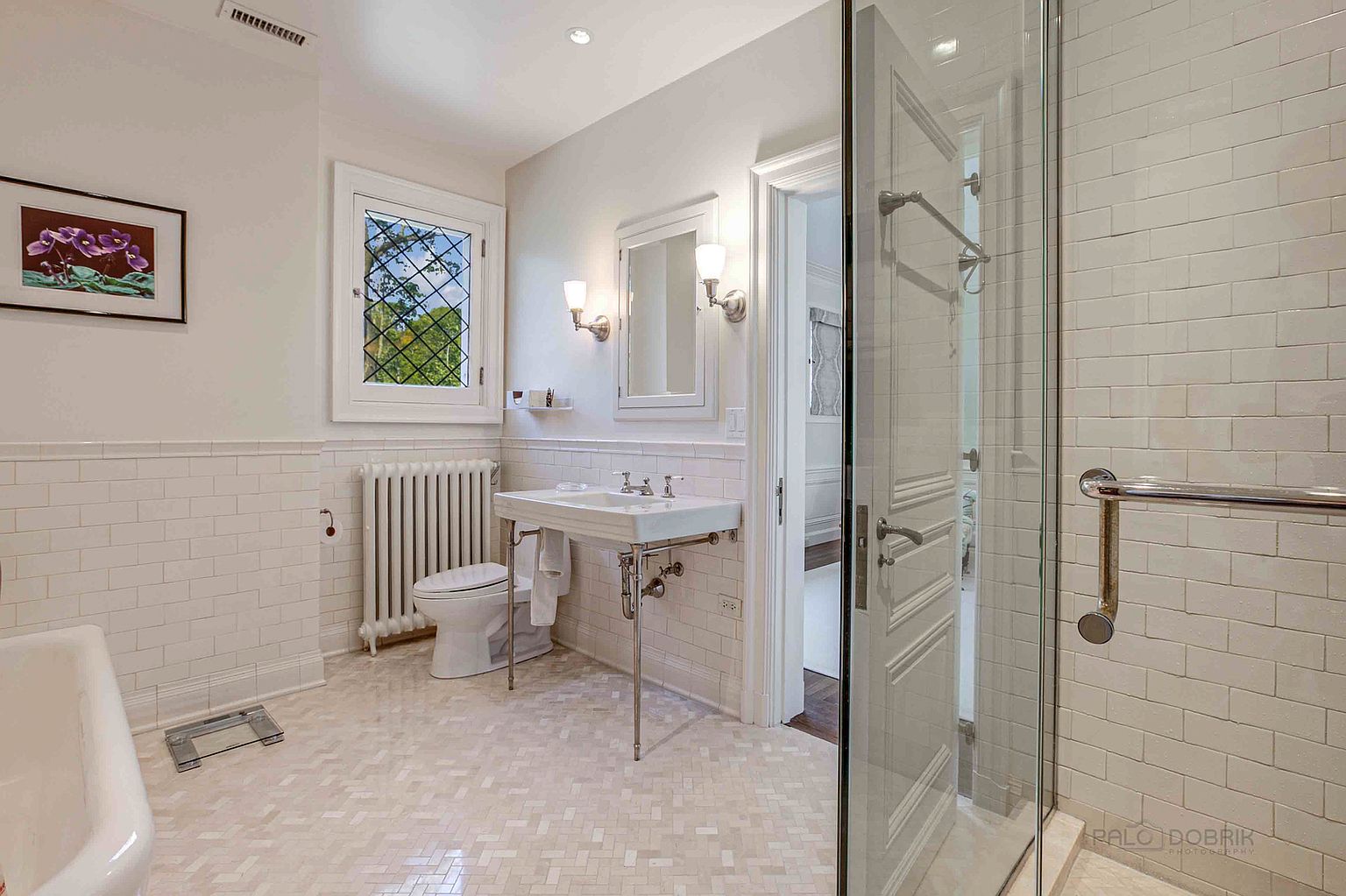 This is a bright and clean primary bathroom featuring white subway tile walls and a mosaic tile floor. The room includes a classic pedestal sink with chrome fixtures, a toilet, a radiator, and a glass-enclosed shower. A window with a decorative lattice provides natural light, and a framed artwork adds a touch of color.