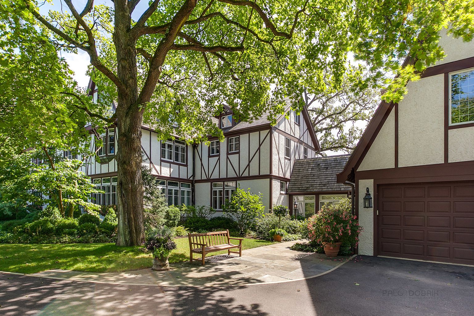 This is a front view of a charming Tudor-style home with a well-manicured lawn and mature trees providing ample shade. The house features a combination of white stucco and dark brown trim, with multiple windows and a visible garage on the right side. A stone pathway leads to a wooden bench, enhancing the inviting curb appeal of the property.