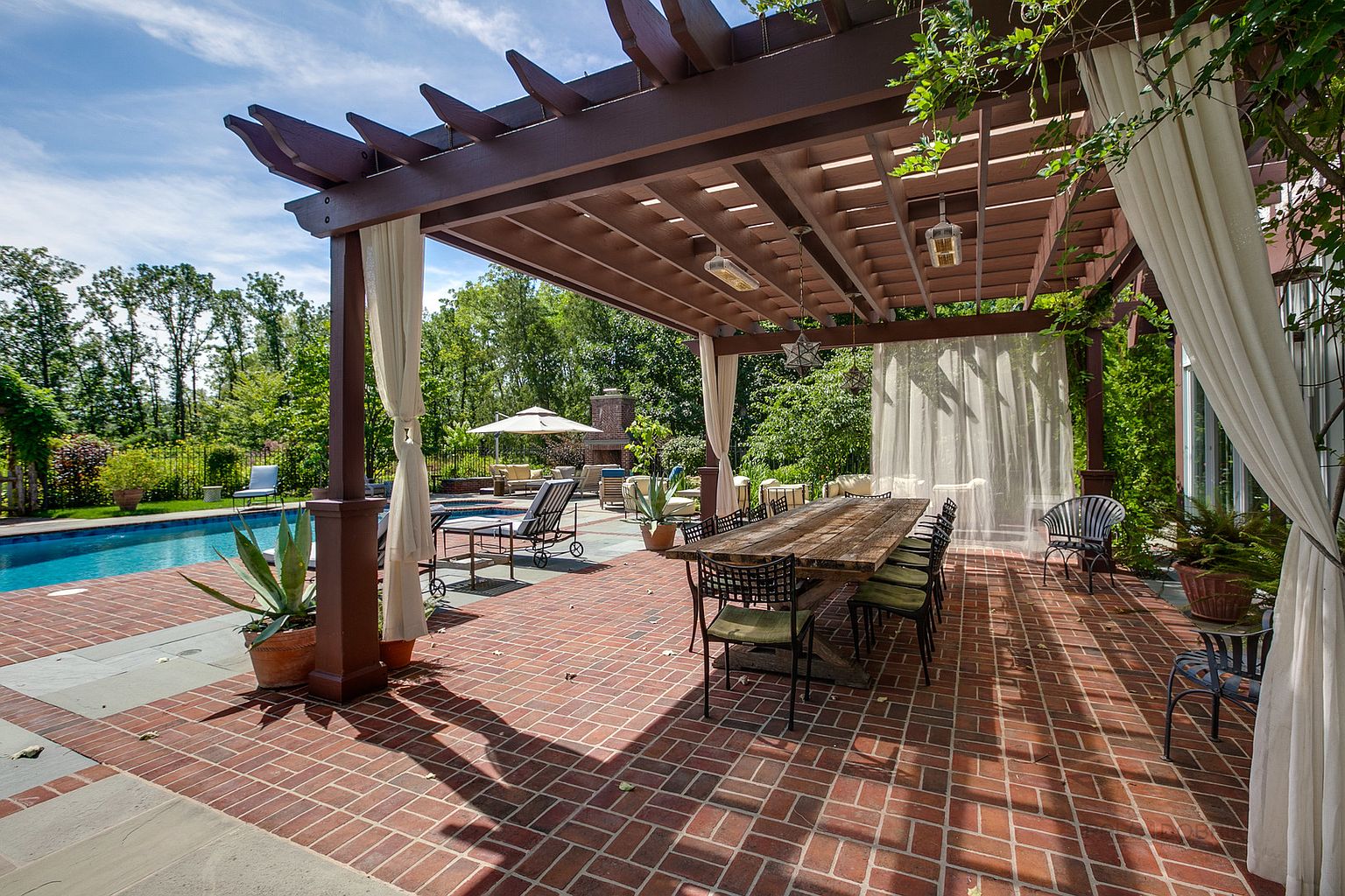 This image showcases a beautifully designed outdoor patio area, featuring a large wooden pergola draped with elegant white curtains. A long wooden dining table with chairs sits on a brick-paved patio, adjacent to a swimming pool and lush greenery. The scene exudes a sense of luxury and relaxation, perfect for outdoor entertaining.