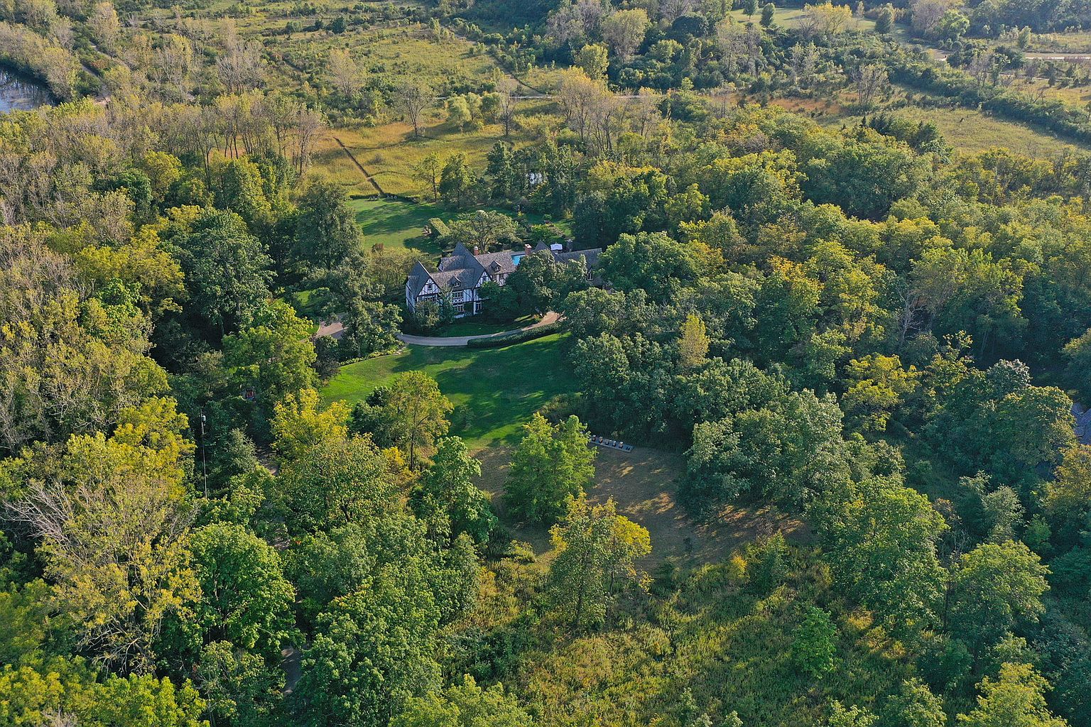 This aerial shot showcases a large, elegant home nestled among lush greenery. The property features a well-manicured lawn, mature trees, and a winding driveway leading to the residence. The architectural style appears to be a Tudor revival with dark wood trim and a steeply pitched roof, creating a sense of timeless charm and privacy.