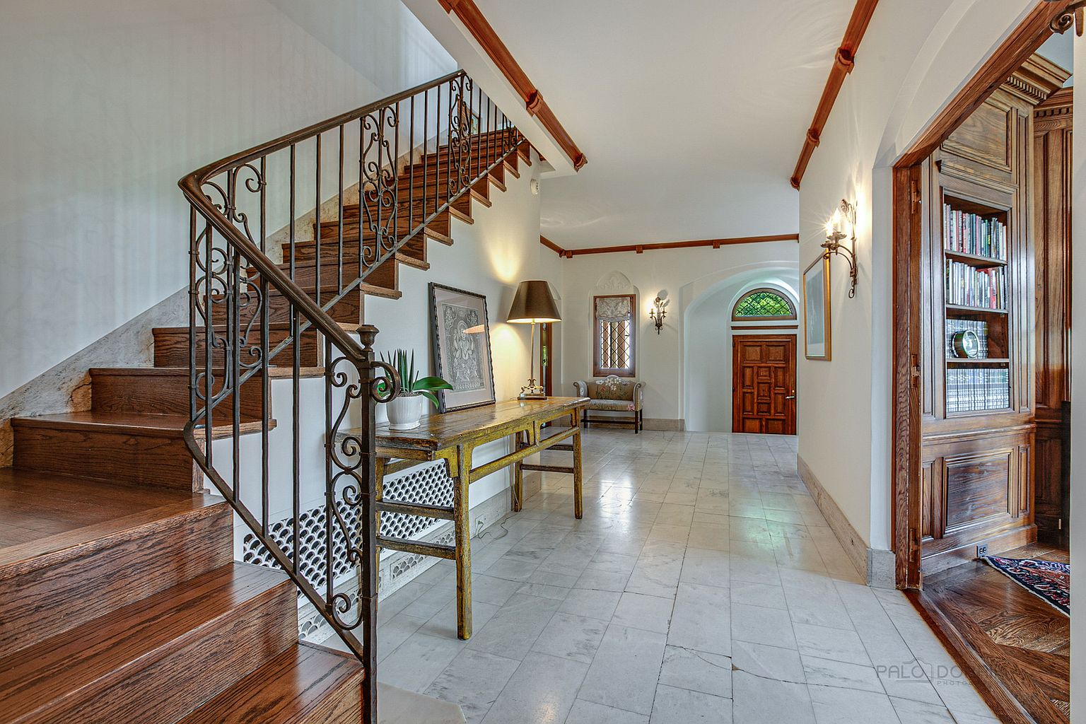 This interior shot showcases a grand hallway and staircase. The staircase features ornate iron railings and wooden steps, while the hallway boasts marble flooring, wooden accents on the ceiling, and a decorative table with a lamp. A doorway to a wood-paneled room with bookshelves is visible on the right, and a front door is visible in the distance, creating a luxurious and inviting atmosphere.