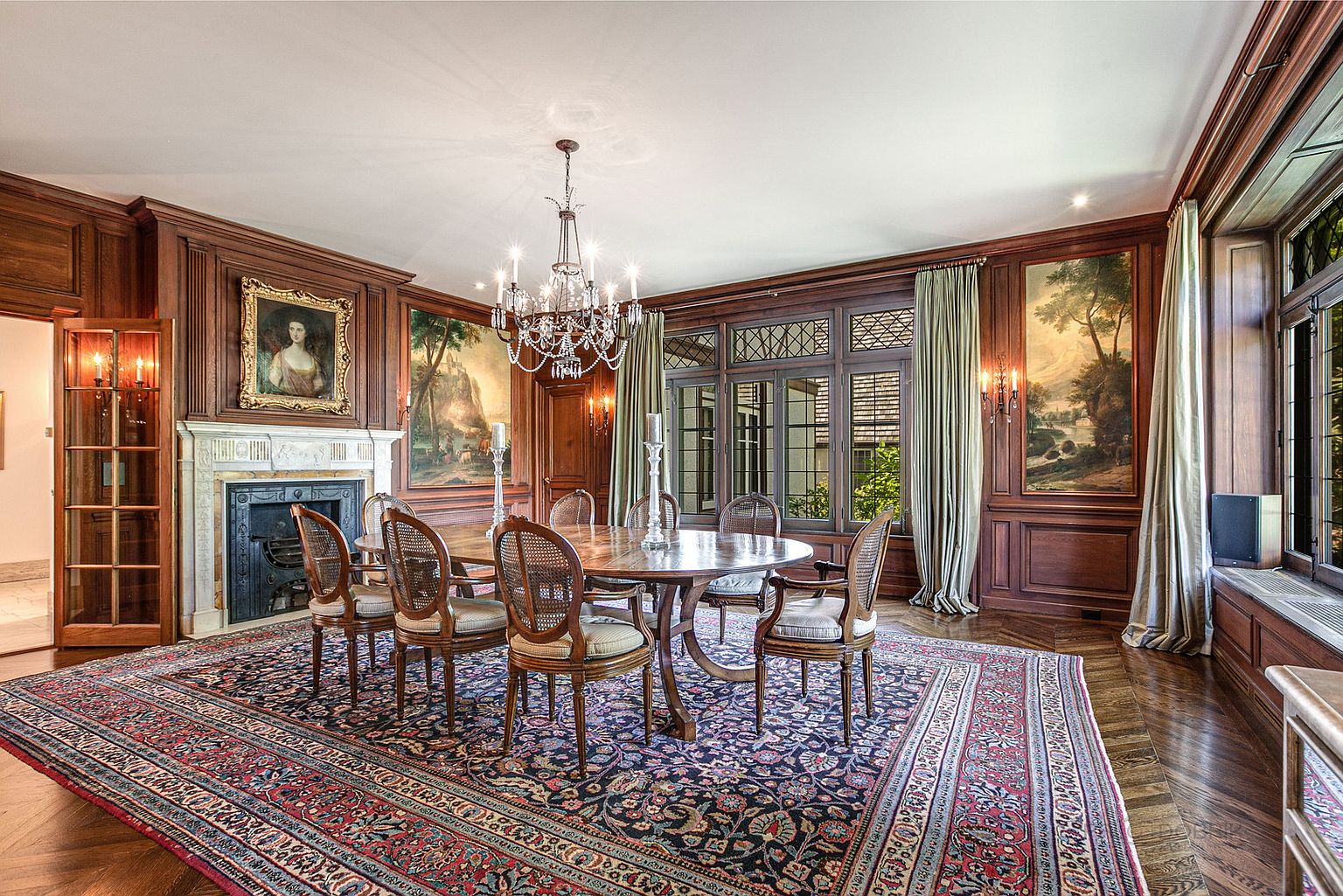 This is a formal dining room featuring dark wood paneling, a large oriental rug, and a round dining table with upholstered chairs. A chandelier hangs above the table, and artwork adorns the walls, creating a sophisticated and elegant atmosphere. The room is well-lit and appears spacious, perfect for entertaining.