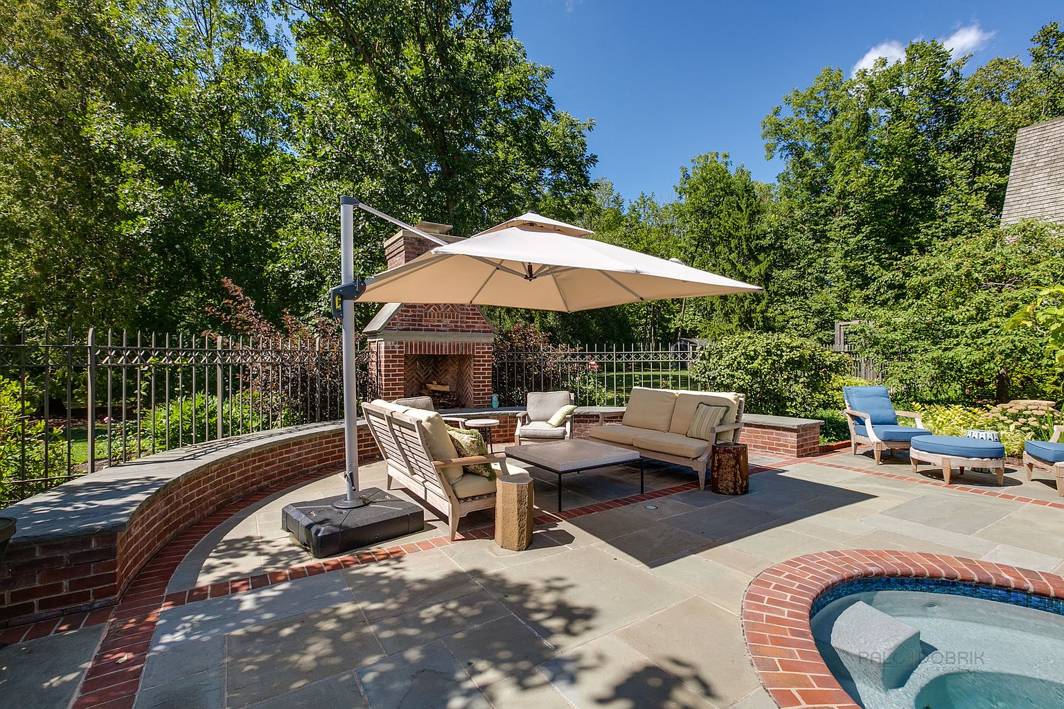 This is a beautifully designed outdoor patio area, featuring comfortable seating arrangements around a coffee table and a brick fireplace. A large umbrella provides shade, and a spa is visible in the corner. The patio is surrounded by lush greenery and a decorative fence, creating a private and inviting outdoor living space.
