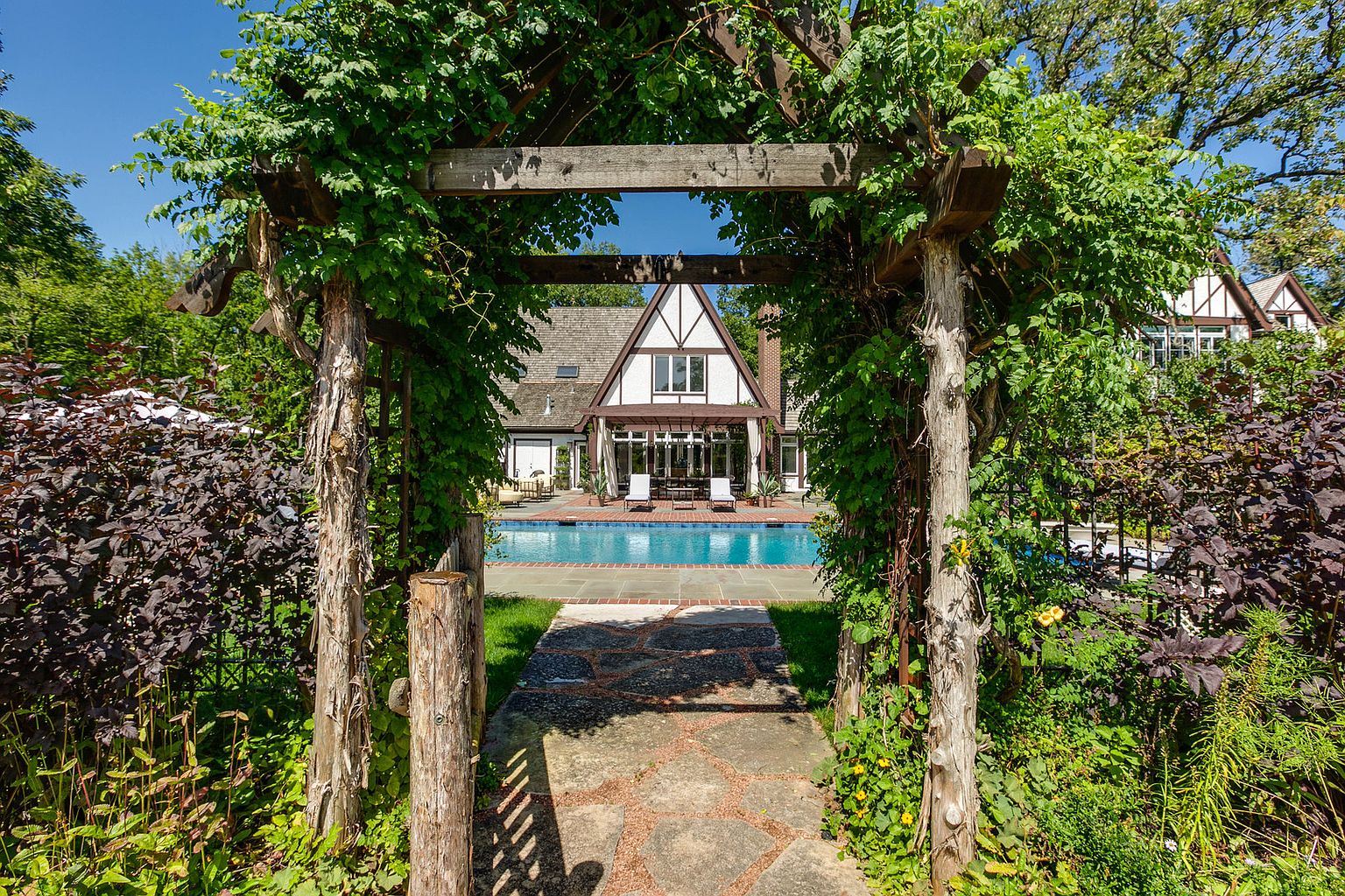 This image showcases a beautiful backyard pool area, viewed through a vine-covered wooden pergola. The pool is surrounded by a brick patio and lush landscaping, leading up to a Tudor-style home with a covered patio area. The scene evokes a sense of tranquility and luxury, perfect for outdoor entertaining and relaxation.