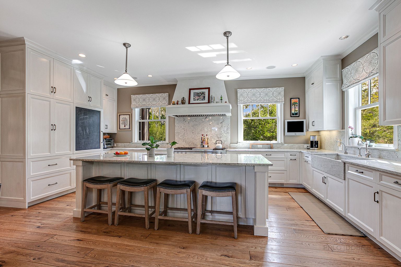 This is a bright and spacious kitchen featuring white cabinetry, granite countertops, and hardwood flooring. A large center island with seating provides ample workspace and a gathering spot. Natural light floods the room through multiple windows, creating a warm and inviting atmosphere.