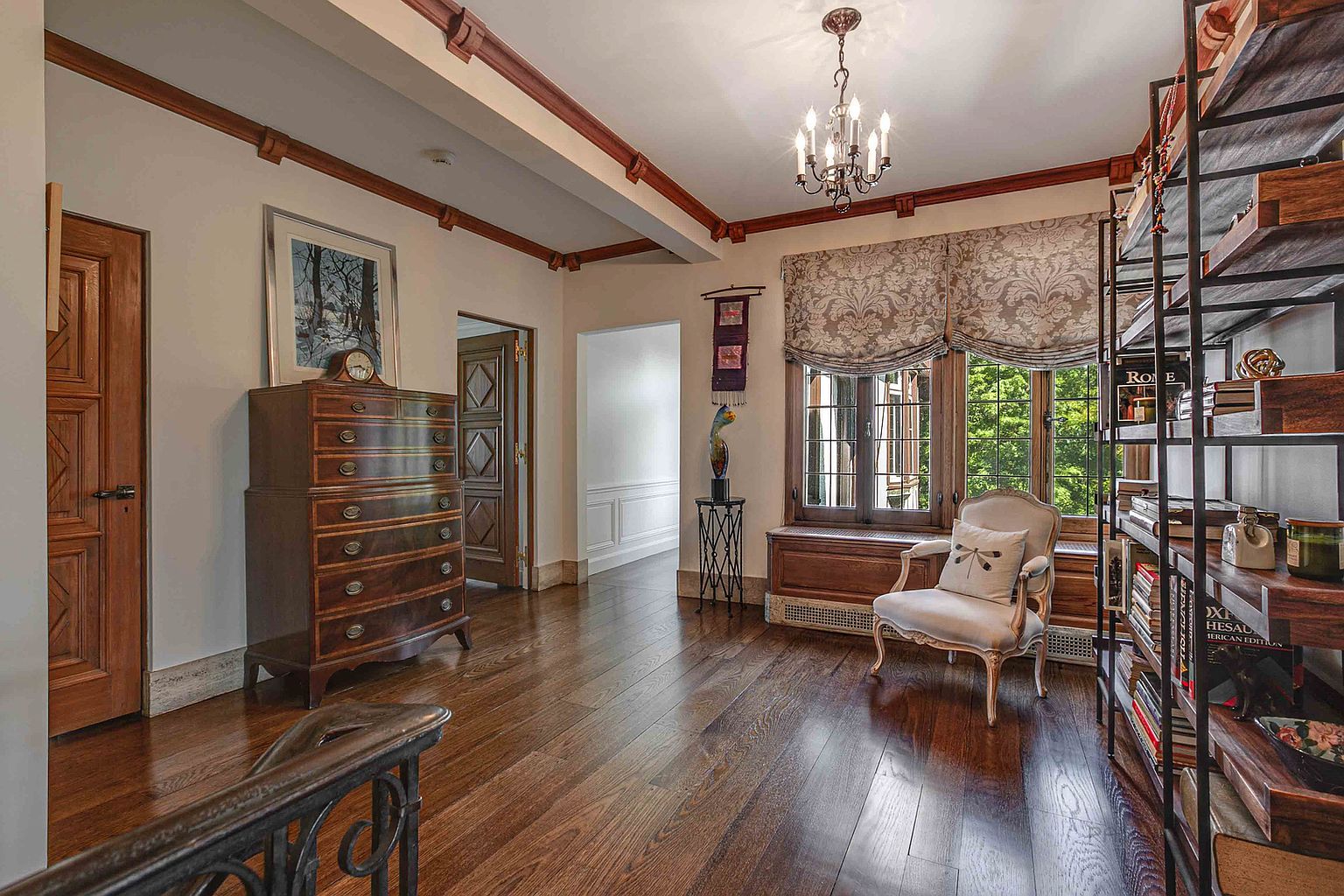 This interior shot showcases a hallway with rich hardwood flooring and elegant architectural details. A dark wood dresser sits against the wall, complemented by a framed artwork above. Natural light streams in through a window adorned with a patterned valance, illuminating a cozy reading nook with an armchair and a bookshelf.