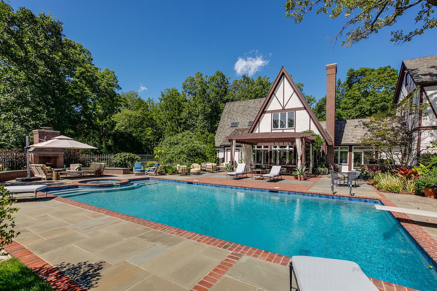 This image showcases a luxurious backyard pool area, complete with a large rectangular pool featuring a brick border and stone patio. The pool is surrounded by various seating arrangements, including lounge chairs and an outdoor fireplace area. The backdrop includes a Tudor-style house with a distinctive chimney and lush greenery, creating a private and upscale outdoor living space.