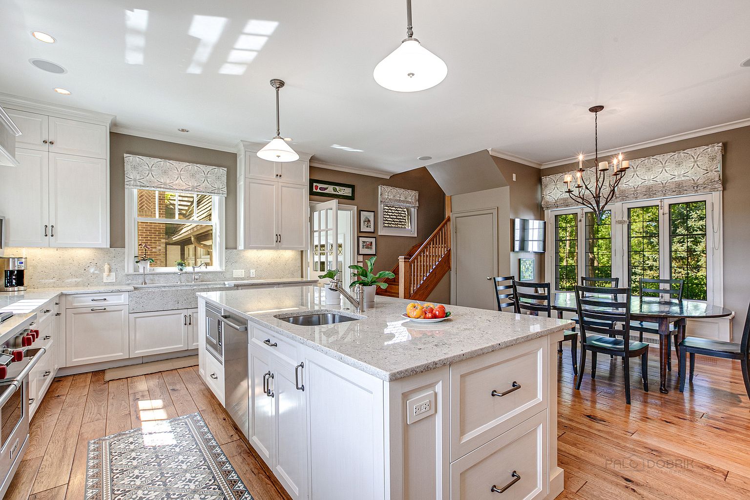 This is a bright and airy kitchen featuring white cabinetry, granite countertops, and stainless steel appliances. A large island provides ample workspace and seating, while pendant lights illuminate the area. The kitchen seamlessly flows into a dining area with a dark wood table and chairs, creating an inviting and functional space.