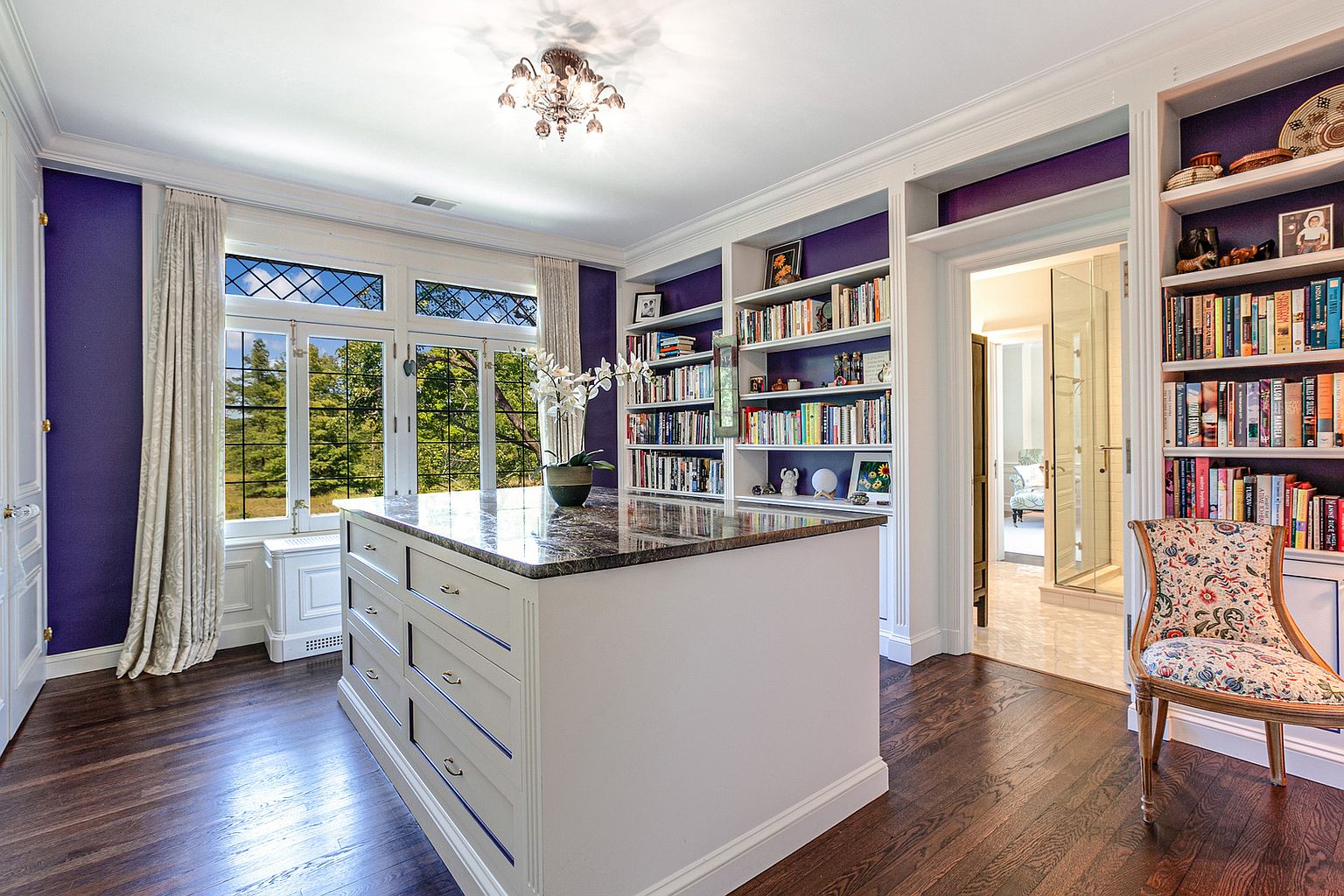 This is an interior shot of a home office or study, featuring dark hardwood floors, purple accent walls, and built-in bookshelves filled with books and decorative items. A large island with drawers sits in the center of the room, and a window provides natural light. The room exudes a sophisticated and organized atmosphere, perfect for a home office or library.