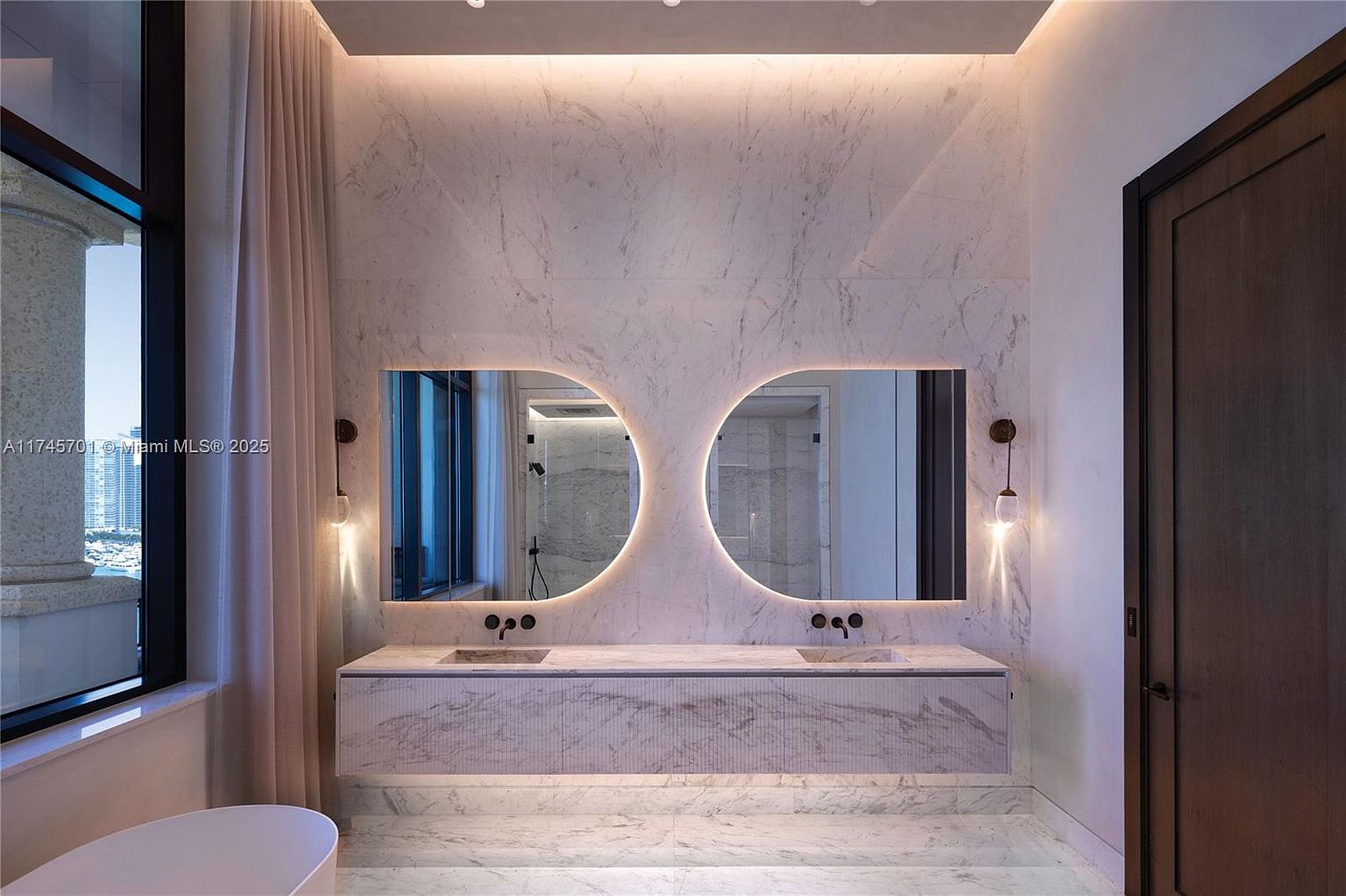 This is a luxurious primary bathroom featuring a double vanity with unique, conjoined circular mirrors illuminated by soft lighting. The walls and floor are covered in marble, creating a seamless and elegant look. A window to the left offers a view of the city, and a door is visible on the right.