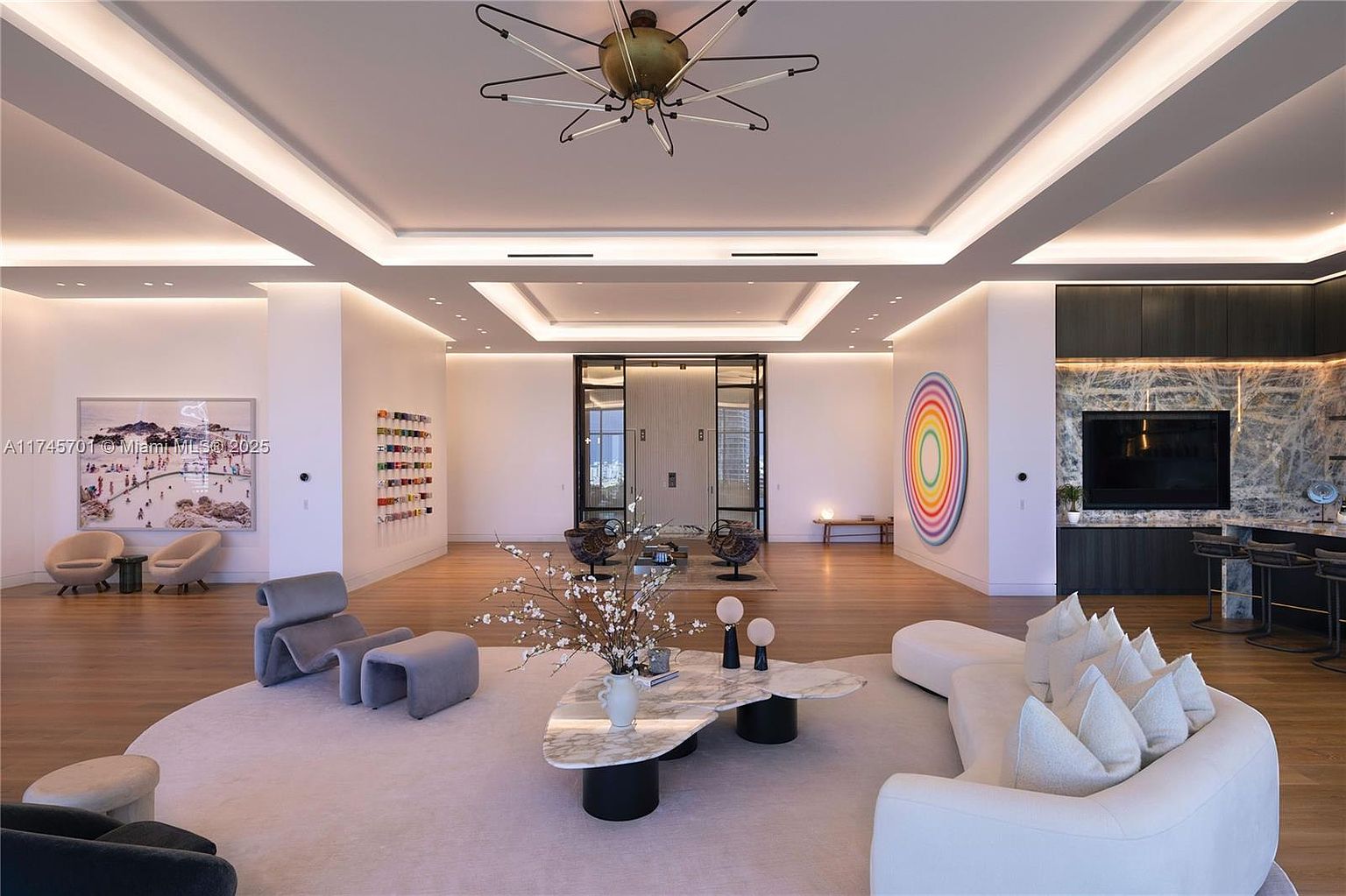 This is a spacious and modern living room featuring a neutral color palette with pops of color from artwork. The room is well-lit with recessed lighting and a unique chandelier. The furniture includes a curved sofa, stylish chairs, and a marble coffee table, all arranged on a large area rug, creating a luxurious and inviting atmosphere.