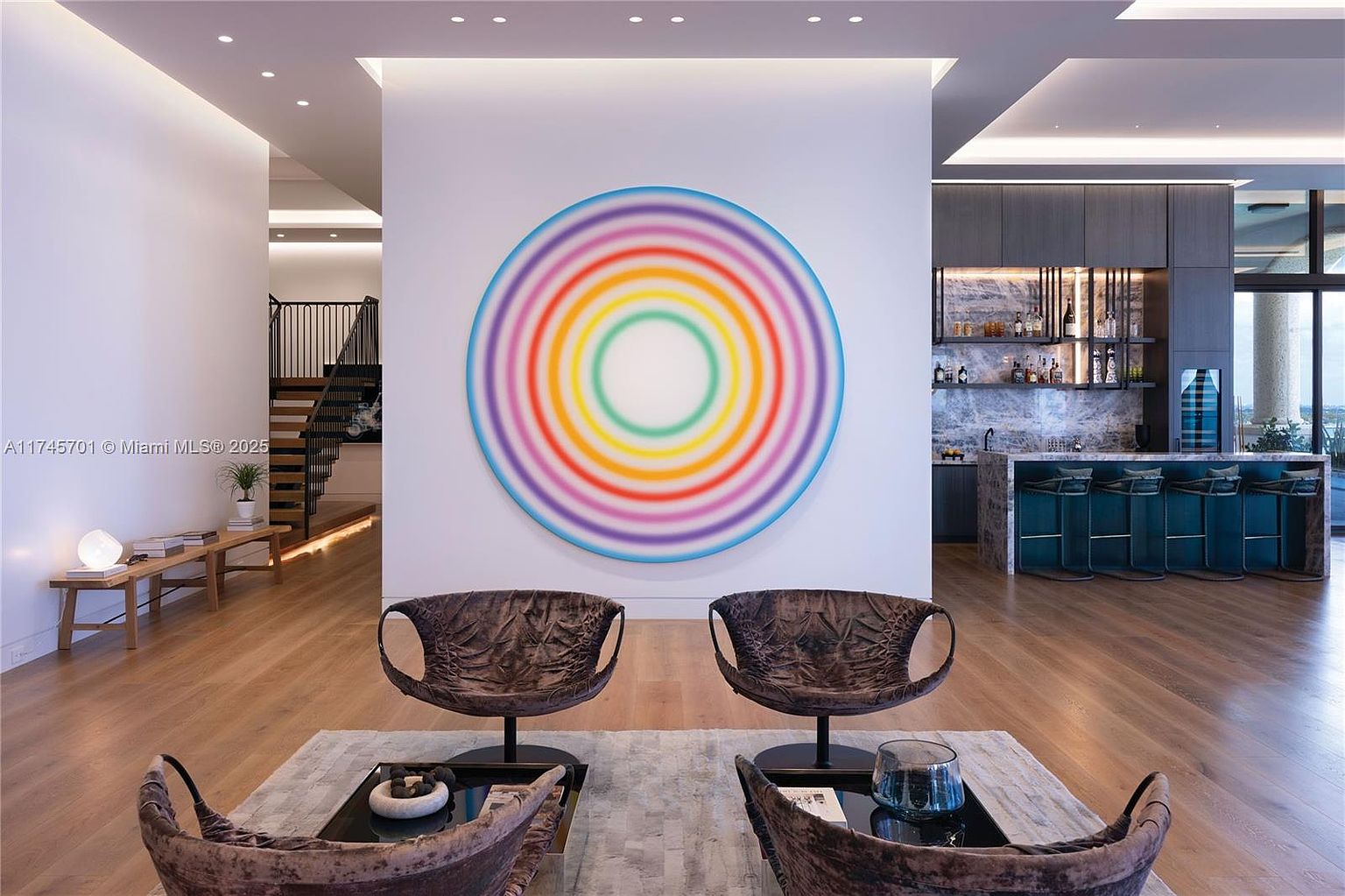 This is an interior shot of a modern living room featuring hardwood floors, a large colorful circular art piece on the wall, and two unique brown armchairs facing the camera. In the background, there's a bar area with stools and a staircase leading to another level, creating a sense of spaciousness and luxury.