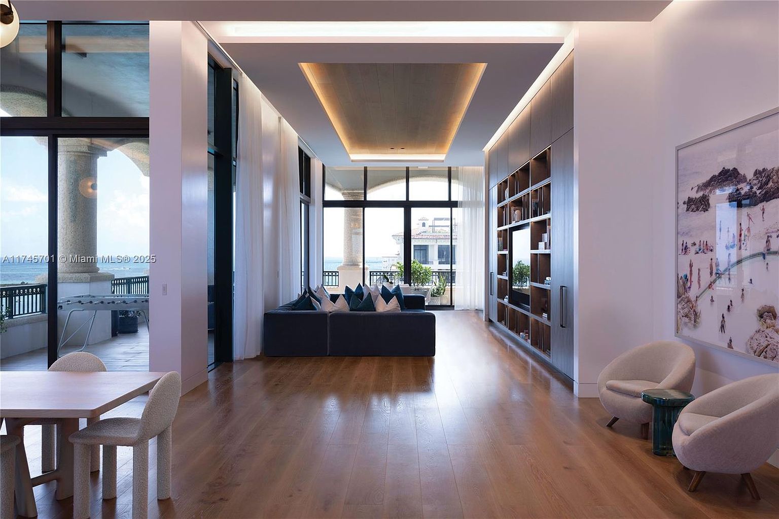 This is a spacious and luxurious living room featuring hardwood floors, a modern blue sofa with decorative pillows, and a custom built-in shelving unit. Large windows offer a view of the ocean and a balcony, while two armchairs and a small table create a cozy seating area. The room is well-lit with recessed lighting and natural light, creating an inviting and elegant atmosphere.