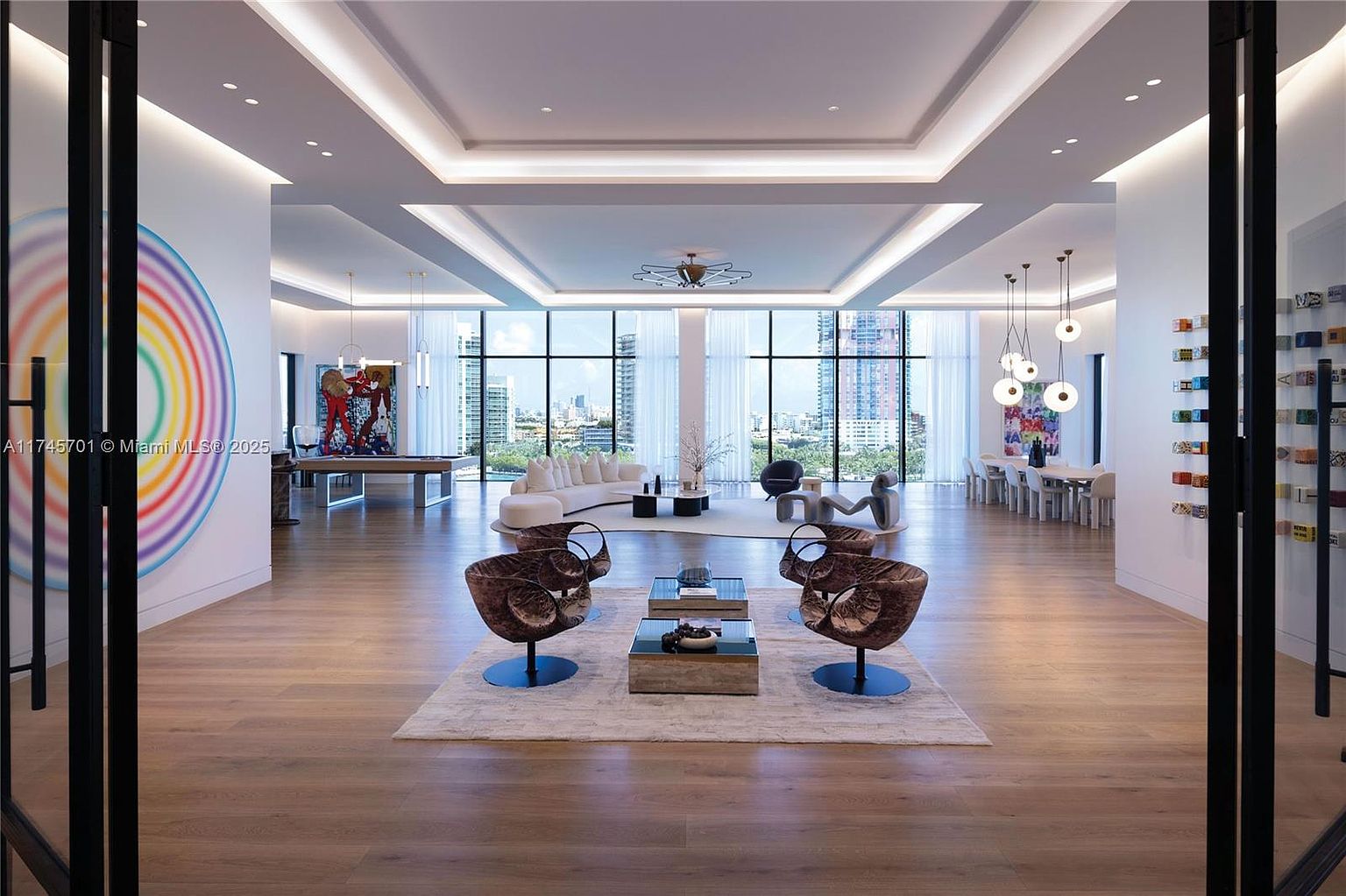 This is a spacious and luxurious living room featuring modern design elements. The room is well-lit with recessed lighting and large windows offering city views. The furniture includes unique chairs, a sleek sofa, and contemporary coffee tables, all arranged on a light-colored rug over hardwood flooring, creating an inviting and stylish atmosphere.