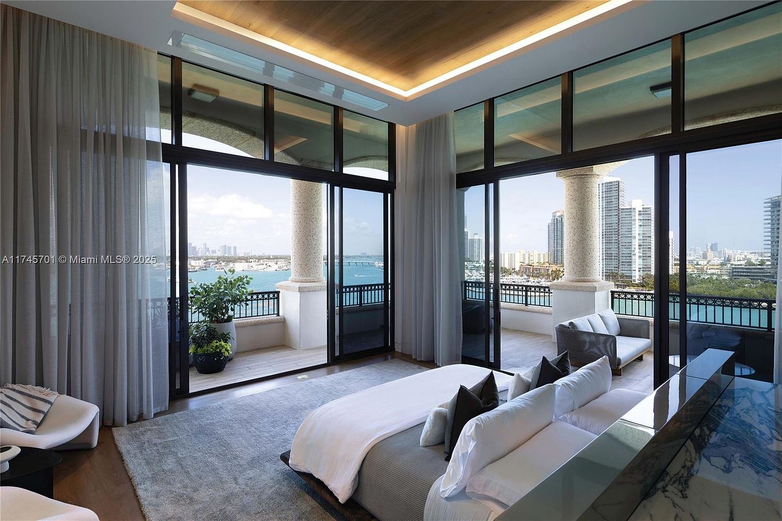 This is a luxurious primary bedroom with floor-to-ceiling windows offering stunning waterfront views. The room features a large bed with plush bedding, a seating area, and access to a balcony. The design is modern and elegant, creating a serene and upscale atmosphere.