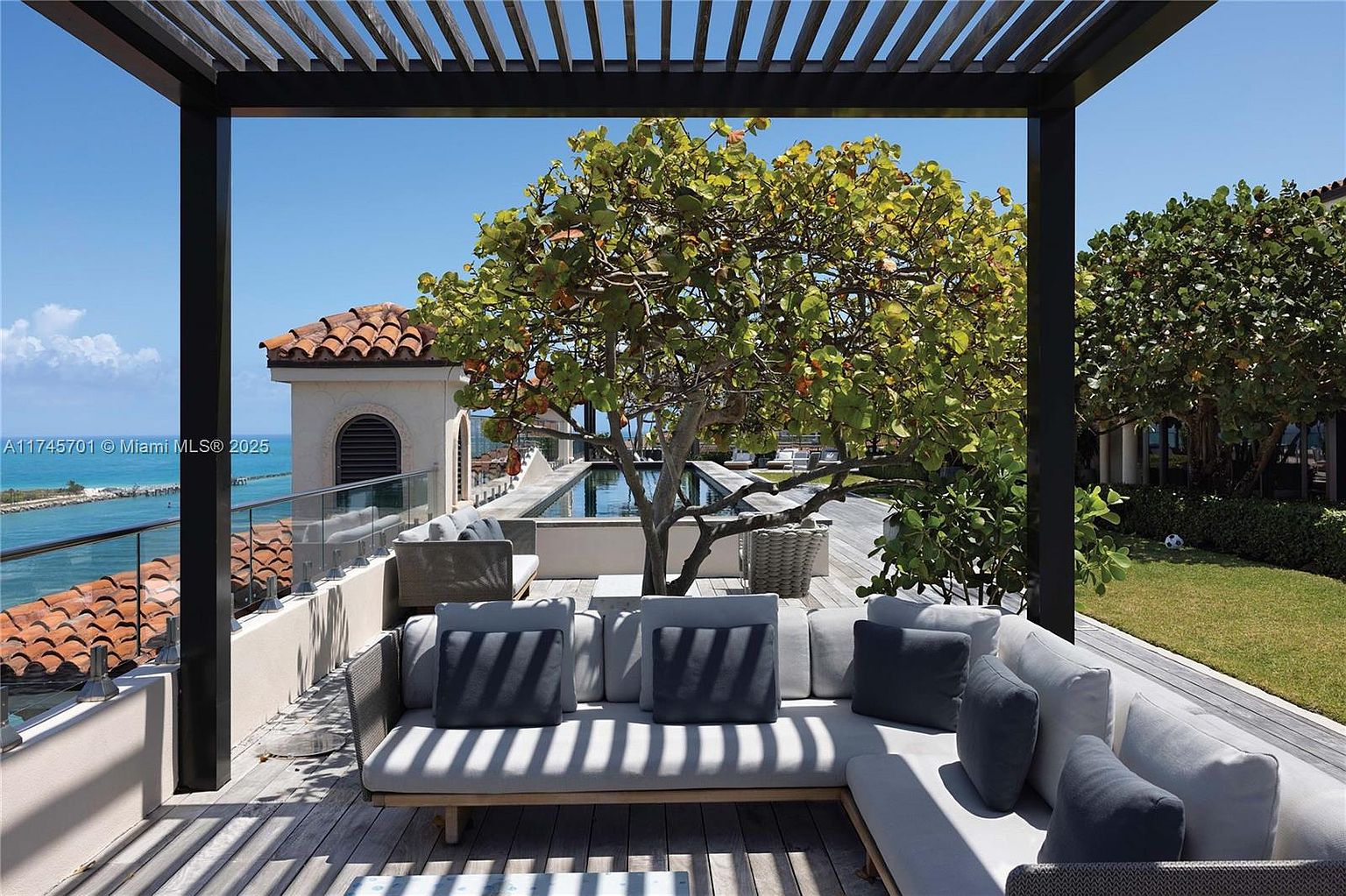 This image showcases a luxurious outdoor patio or deck area, featuring comfortable lounge seating with stylish cushions and pillows. The space is covered by a modern pergola, providing shade and architectural interest. A tree adds a natural element, while the view includes a glimpse of the ocean and a well-maintained lawn, creating an inviting and upscale outdoor living space.