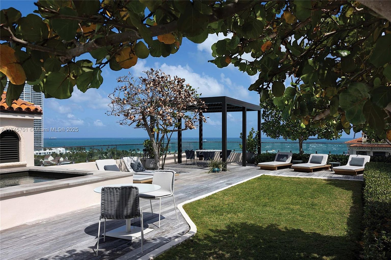 This image showcases a luxurious rooftop patio/deck/balcony with stunning ocean views. The space features a combination of wooden decking, a small patch of green lawn, and modern outdoor furniture including lounge chairs, a dining table, and a pergola. The overall impression is one of high-end outdoor living and relaxation.