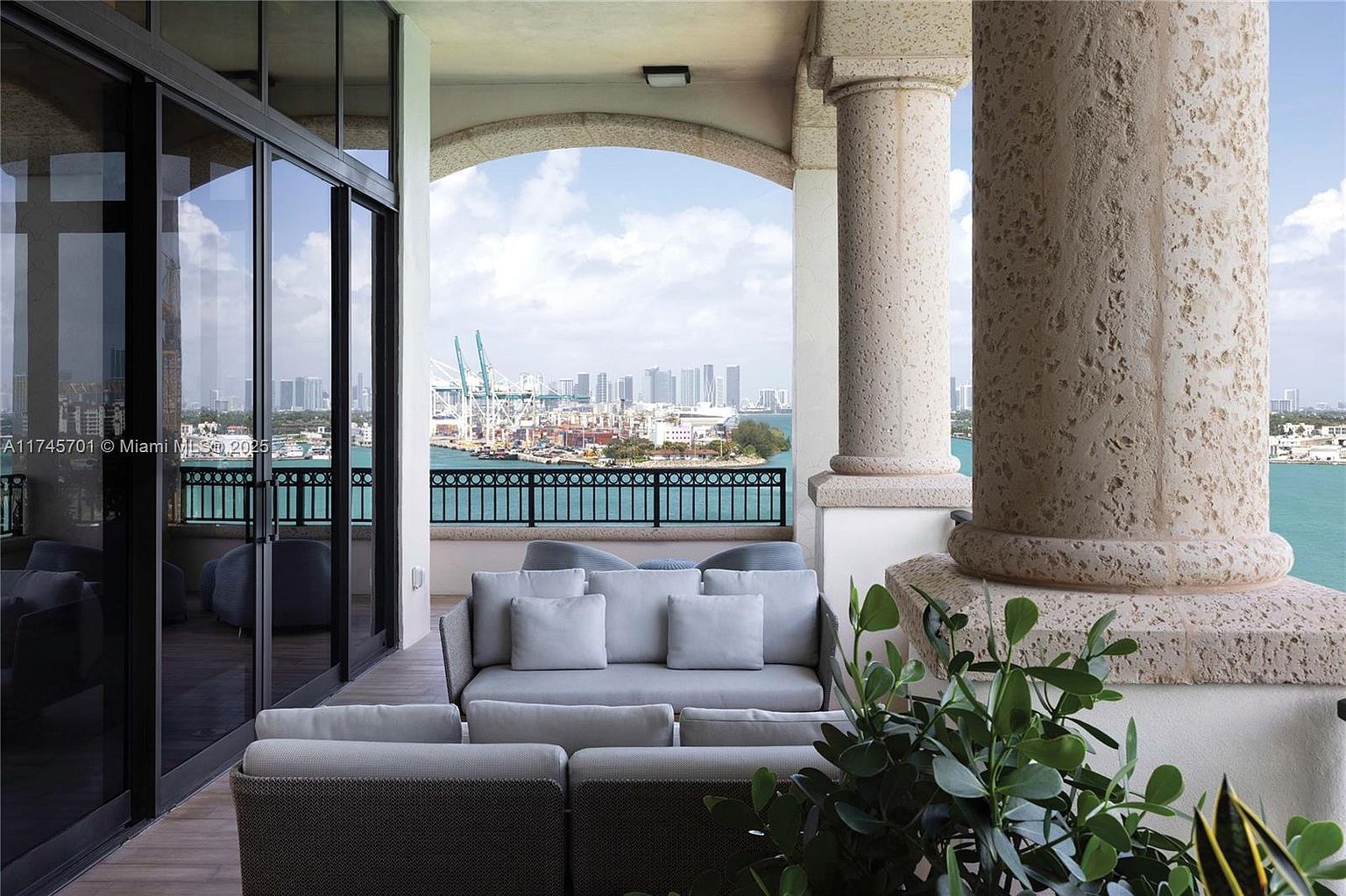 This image showcases a luxurious balcony with comfortable outdoor seating, including a gray sofa and chairs. The balcony features architectural columns and an arched opening that frames a stunning view of the city skyline and waterfront. The space is well-appointed and offers a relaxing outdoor living area.