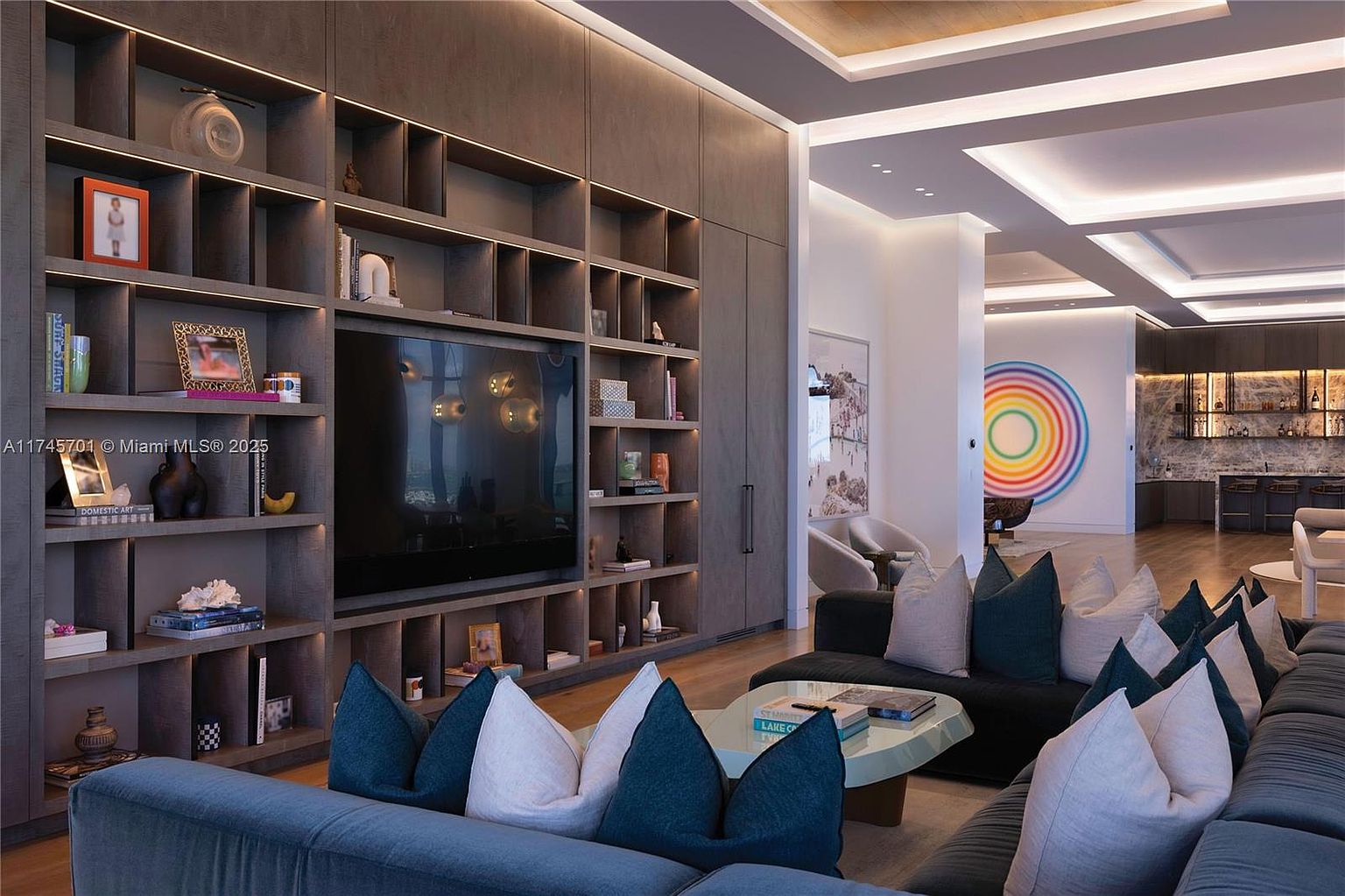 This is an interior shot of a modern living room featuring a large, blue sectional sofa with decorative pillows. A custom built-in shelving unit with a large television provides a focal point, while the open floor plan leads to a bar area and a colorful art piece. The room is well-lit with recessed lighting and has a sophisticated and inviting atmosphere.