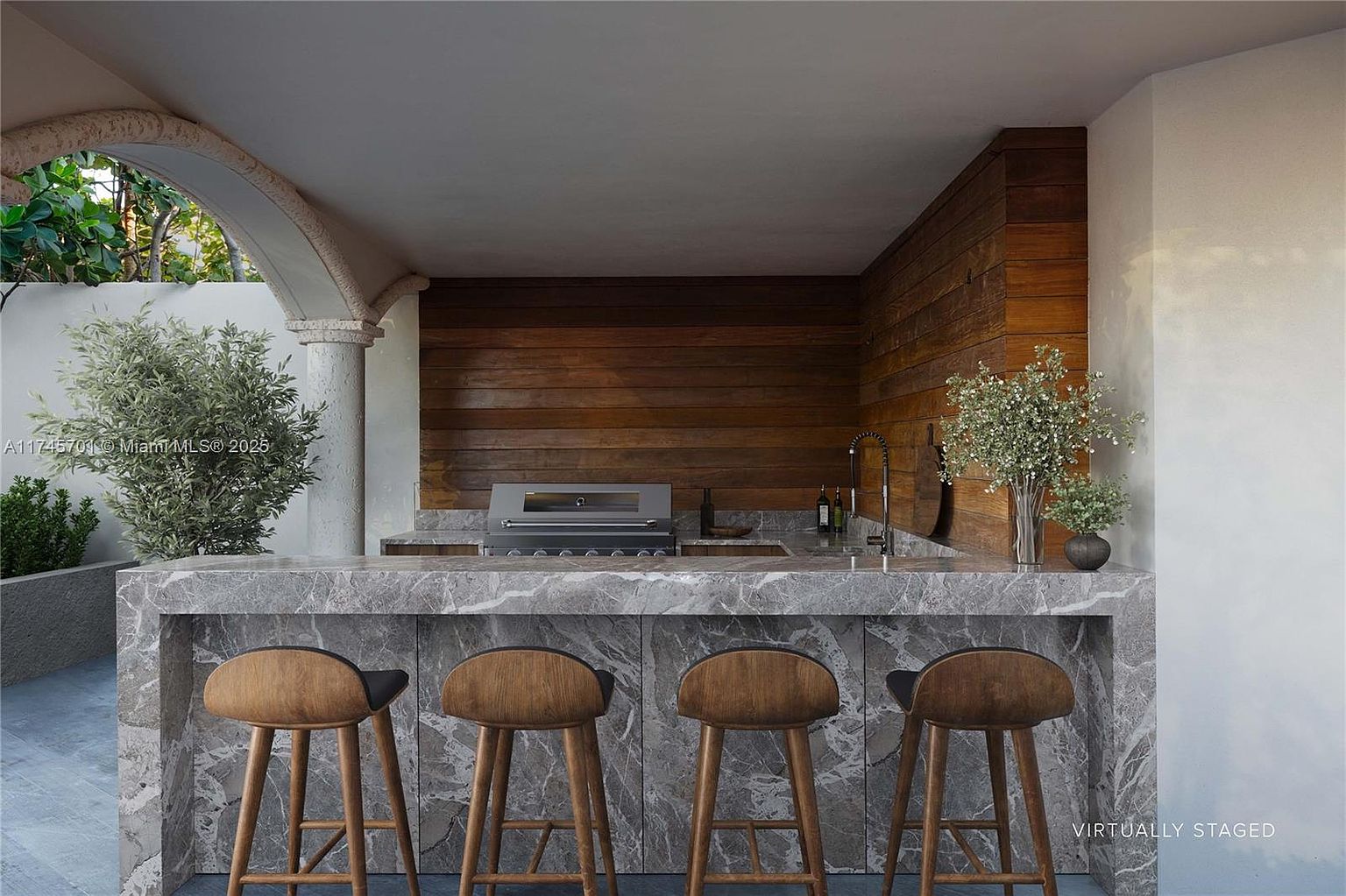 This image showcases an outdoor kitchen area, featuring a marble countertop with bar seating. The kitchen is equipped with a built-in grill and sink, set against a backdrop of wood paneling. The space is partially covered by an arched structure, creating a stylish and functional outdoor living area.