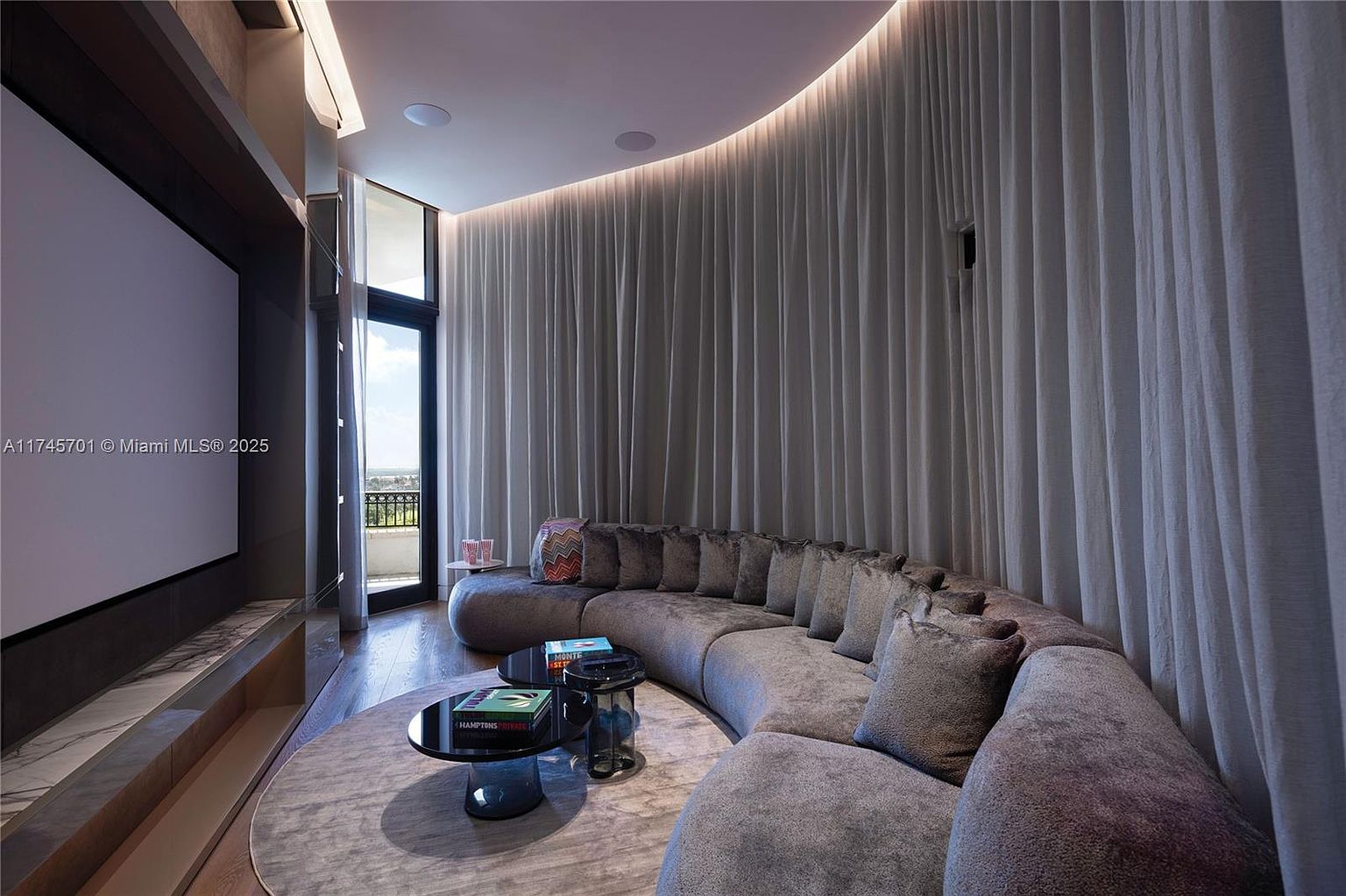 This is an interior shot of a modern living room featuring a large, curved gray sofa with numerous pillows, positioned on a round rug. A black coffee table sits in front of the sofa, and floor-to-ceiling gray curtains line the curved wall behind it, creating a cozy and cinematic atmosphere. A large screen is visible on the left, suggesting a home theater setup.
