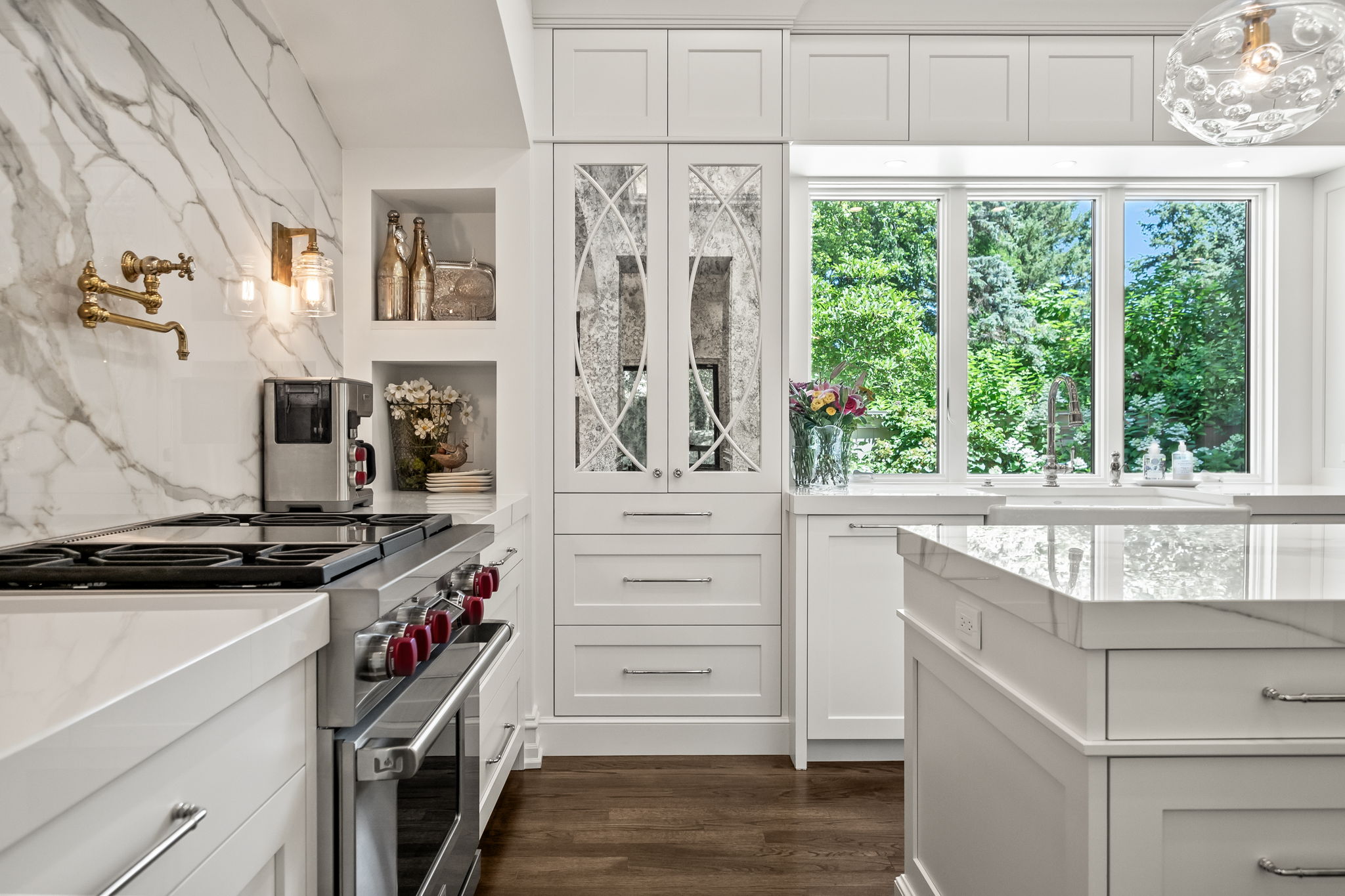 This is a bright and elegant kitchen featuring white cabinetry, marble countertops, and stainless steel appliances. A large window provides natural light and a view of lush greenery. The kitchen also includes decorative mirrored cabinets and a stylish range with red knobs, creating a sophisticated and functional space.