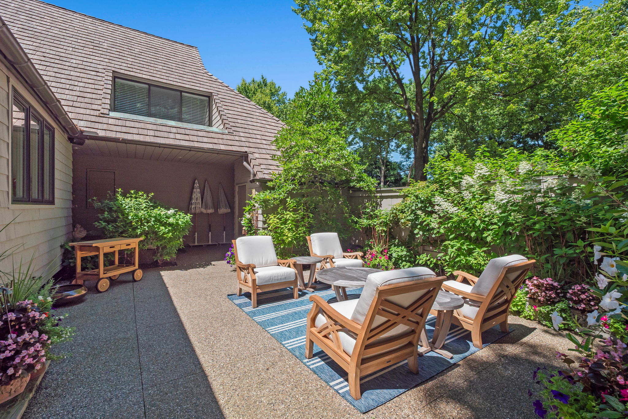 This image showcases a charming outdoor patio area, perfect for relaxation and entertaining. The space is furnished with comfortable wooden chairs and small side tables, arranged around a central coffee table on a blue striped rug. Lush greenery surrounds the patio, creating a private and inviting atmosphere, while the house provides a sheltered backdrop.