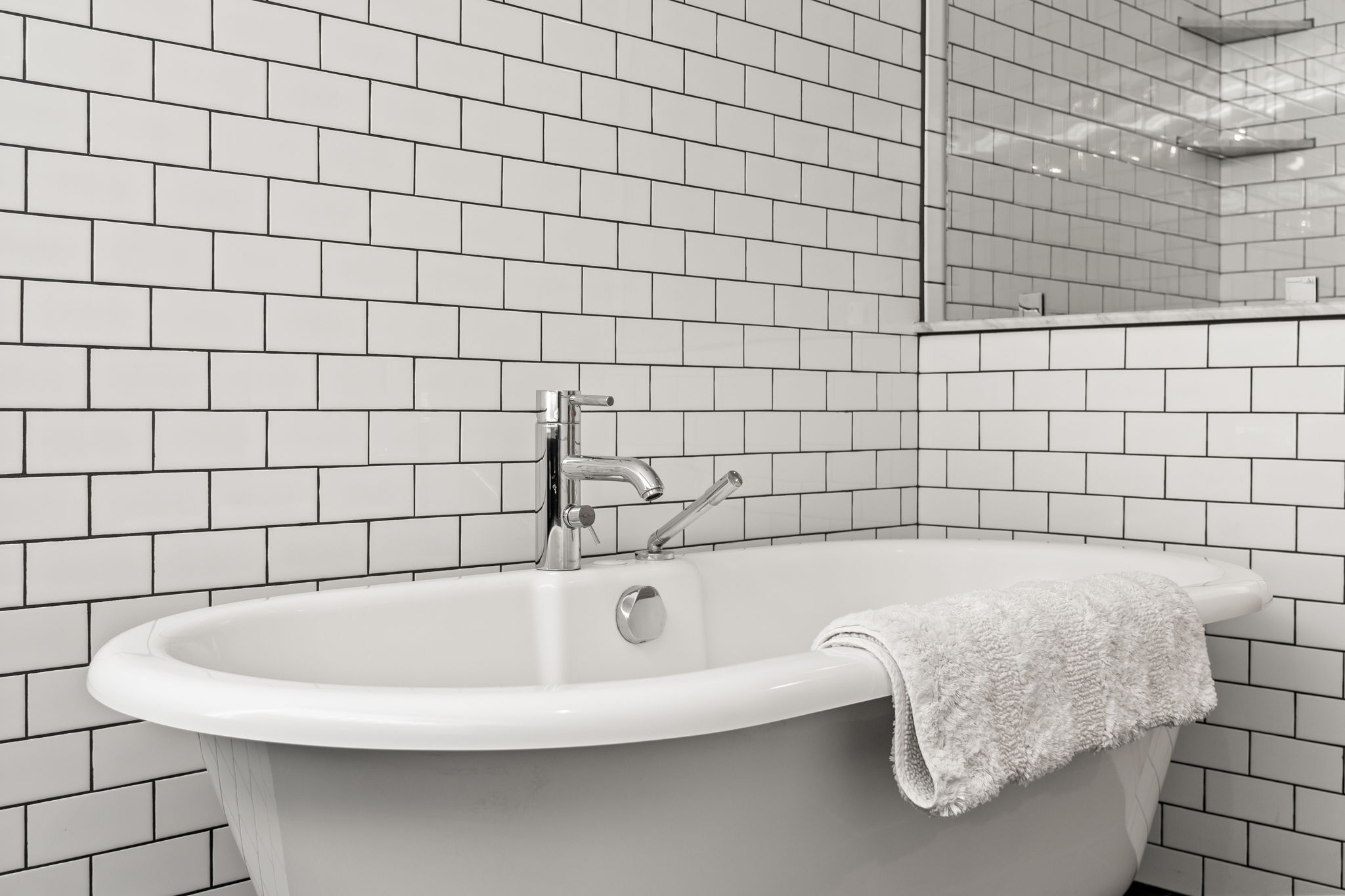 This is a bright and clean primary bathroom featuring a freestanding bathtub with chrome fixtures. The walls are covered in classic white subway tiles with dark grout, creating a modern yet timeless look. A plush white towel is draped over the edge of the tub, adding a touch of luxury.