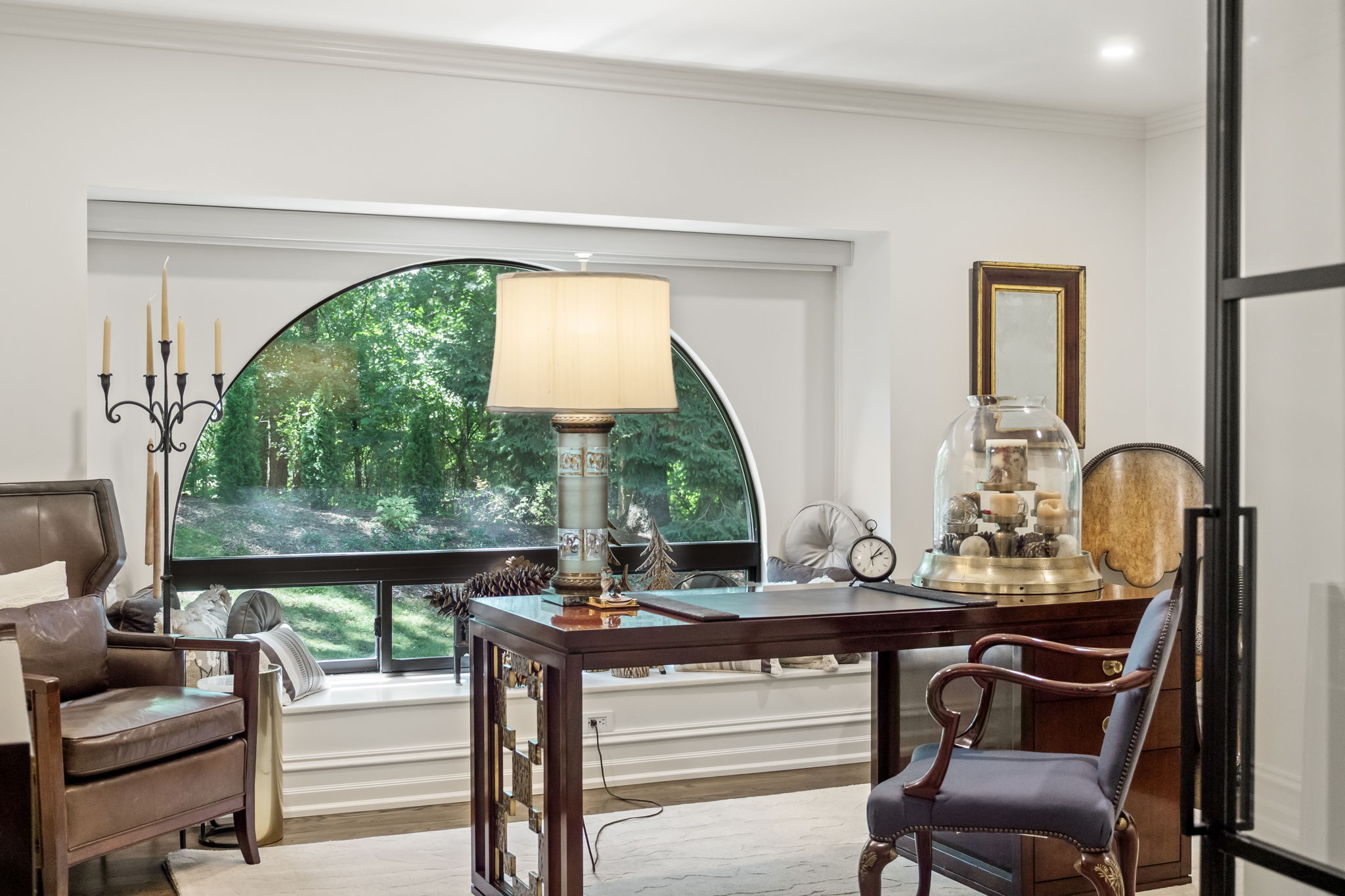 This is an interior shot of a well-appointed office or study. The room features a dark wood desk with a lamp, a comfortable leather chair, and a window offering a view of lush greenery. The overall impression is one of sophistication and tranquility, ideal for a productive workspace.