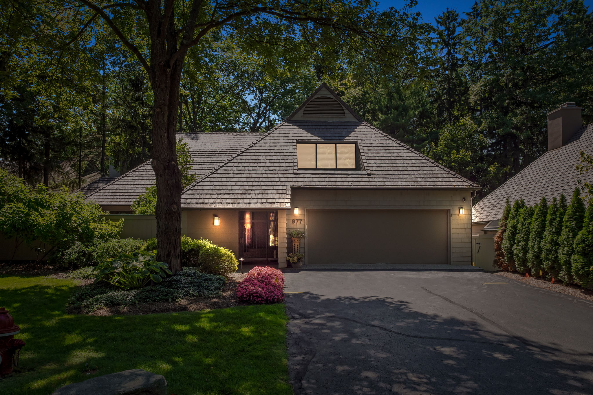 The image showcases the front exterior of a single-story home with a unique architectural style. The house features a brown shingle roof, a two-car garage, and a well-maintained front yard with mature trees and landscaping. The facade is illuminated by warm lighting, creating an inviting and cozy atmosphere.