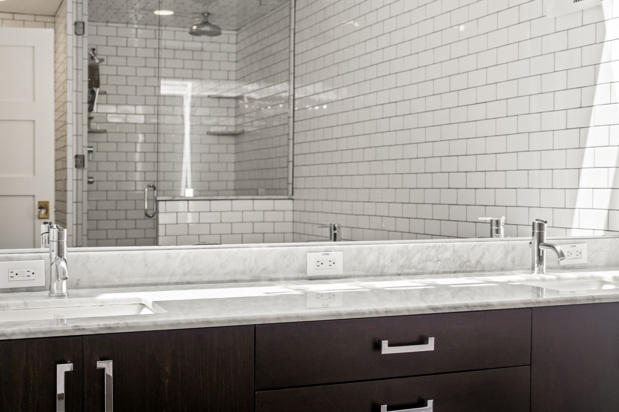 This is a well-lit primary bathroom featuring a double vanity with a marble countertop and dark wood cabinetry. The backsplash and shower are tiled with white subway tiles, creating a clean and modern aesthetic. Chrome fixtures and hardware add a touch of elegance to the space.