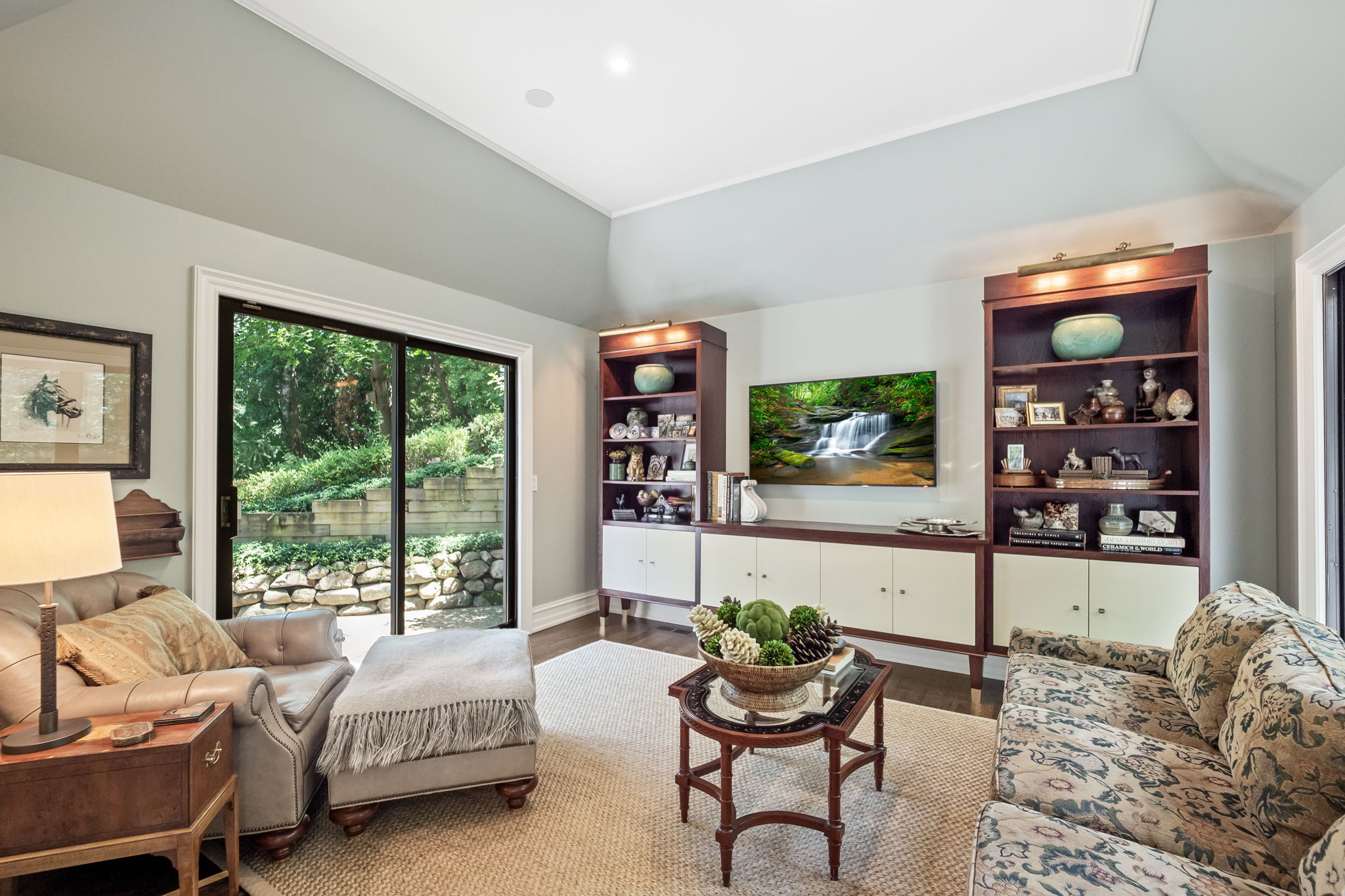This is a well-decorated living room featuring a comfortable armchair, a floral patterned sofa, and built-in shelving units flanking a television. A sliding glass door provides natural light and a view of the outdoor greenery. The room is furnished with a coffee table and ottoman, creating a cozy and inviting atmosphere.