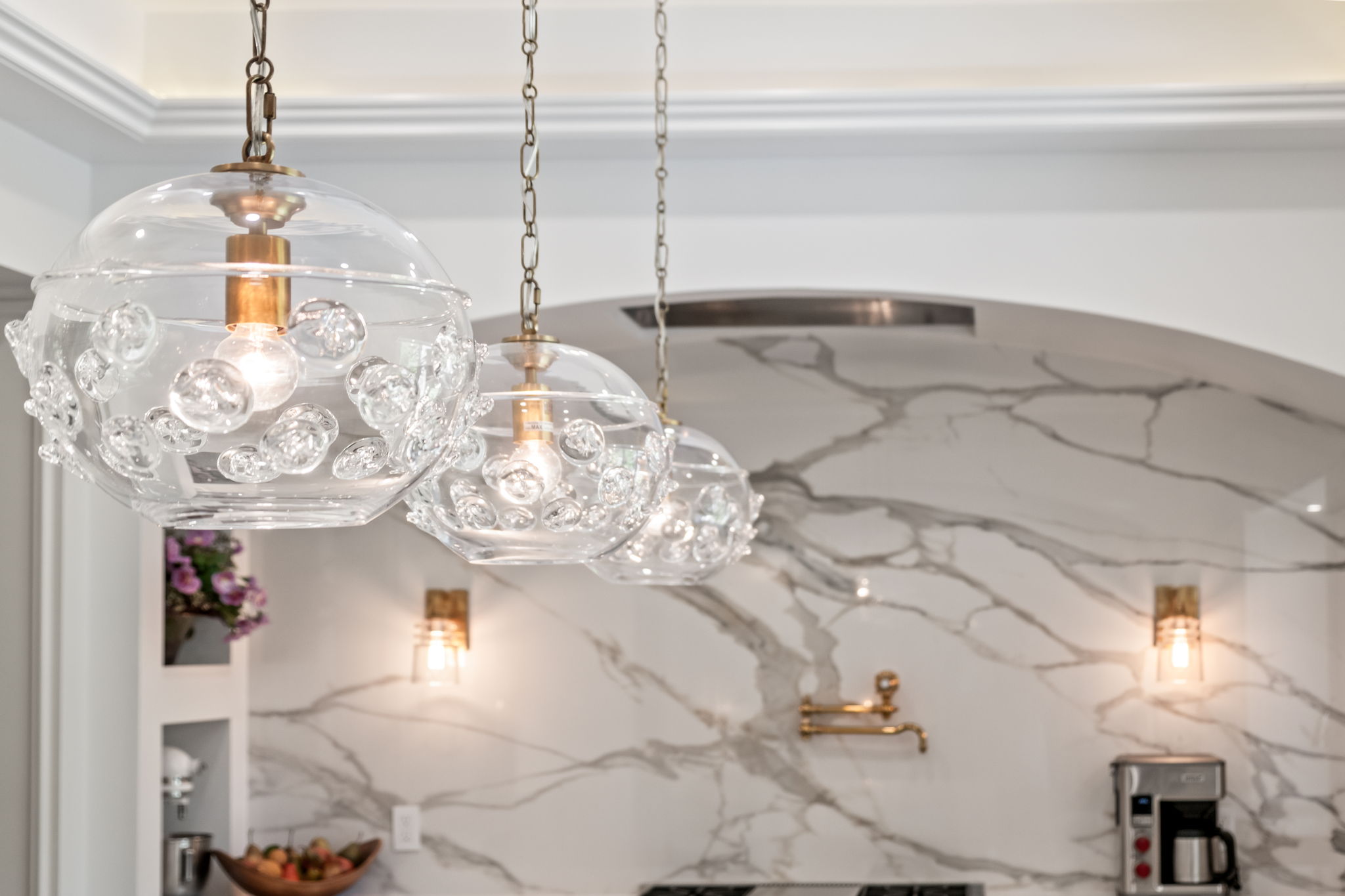 This image showcases a stylish kitchen interior, focusing on three decorative glass pendant lights hanging above the countertop. The backdrop features a marble-patterned wall, adding a touch of luxury, complemented by brass fixtures and sconces. The overall impression is one of modern elegance and sophisticated design.