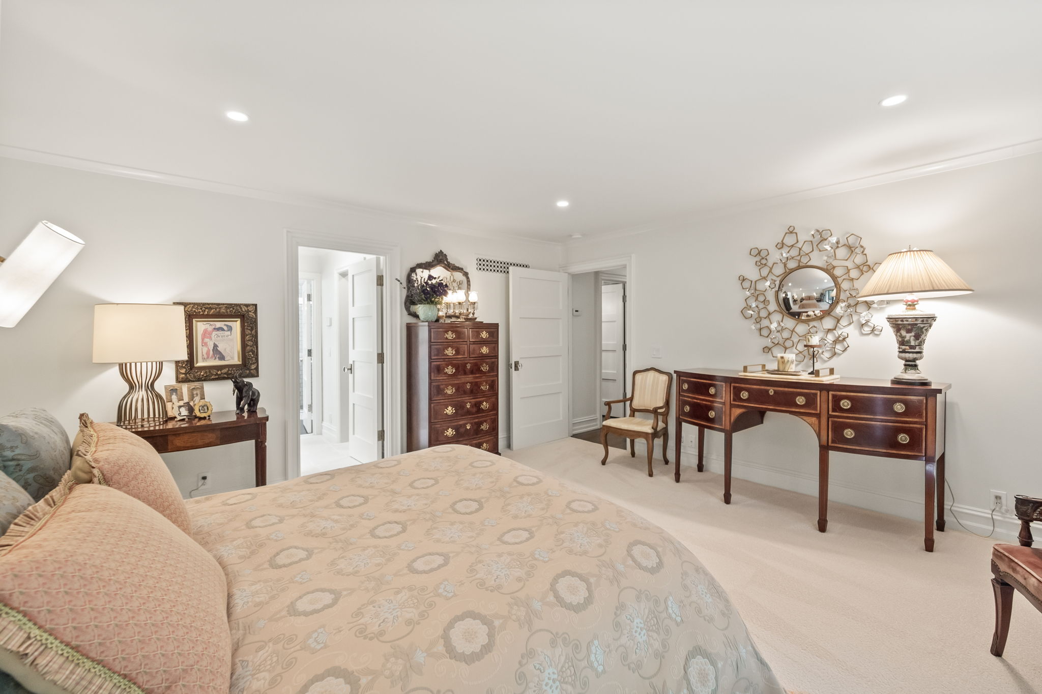 This is a well-lit primary bedroom featuring a large bed with patterned bedding and decorative pillows. The room is furnished with antique-style wooden furniture, including a dresser, desk, and side table, complemented by ornate mirrors and lamps. The overall impression is elegant and traditional, with a focus on comfort and classic design.