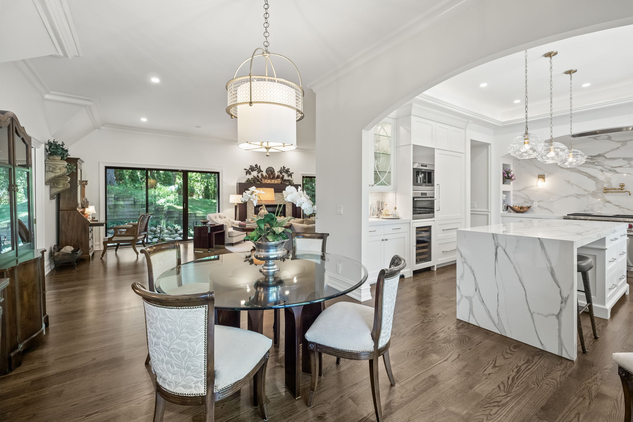 This interior shot showcases a dining area adjacent to a modern kitchen and a living room. The dining area features a round glass table with upholstered chairs beneath a decorative chandelier. The kitchen boasts white cabinetry, a marble island, and pendant lighting, while the living room offers a view of the outdoors through large windows, creating an open and inviting atmosphere.