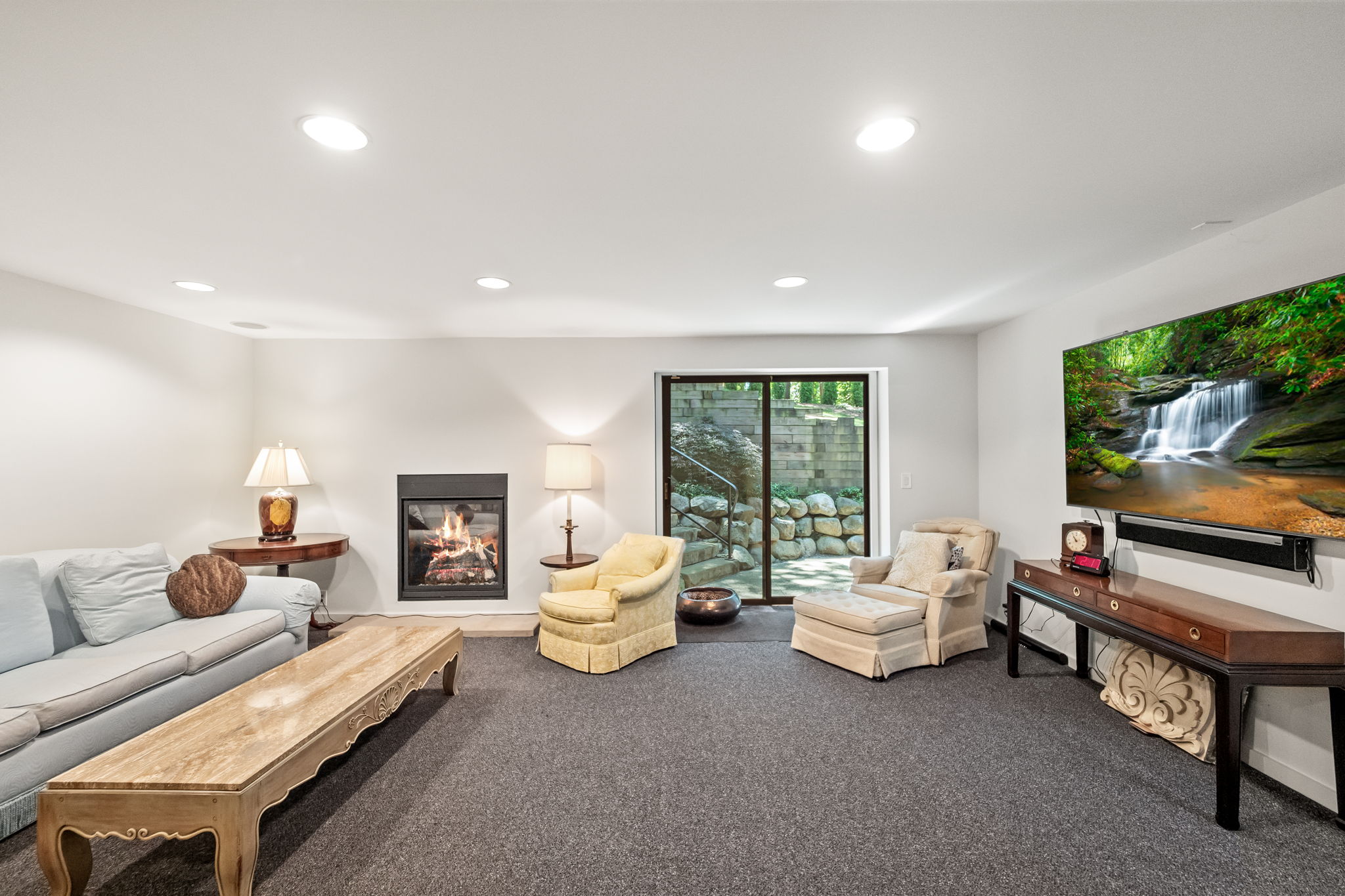 This is a cozy living room featuring a fireplace, a comfortable light blue sofa, and two armchairs. A large television is mounted on the wall above a console table. The room has neutral-colored walls and carpet, creating a warm and inviting atmosphere, perfect for relaxation and entertainment.