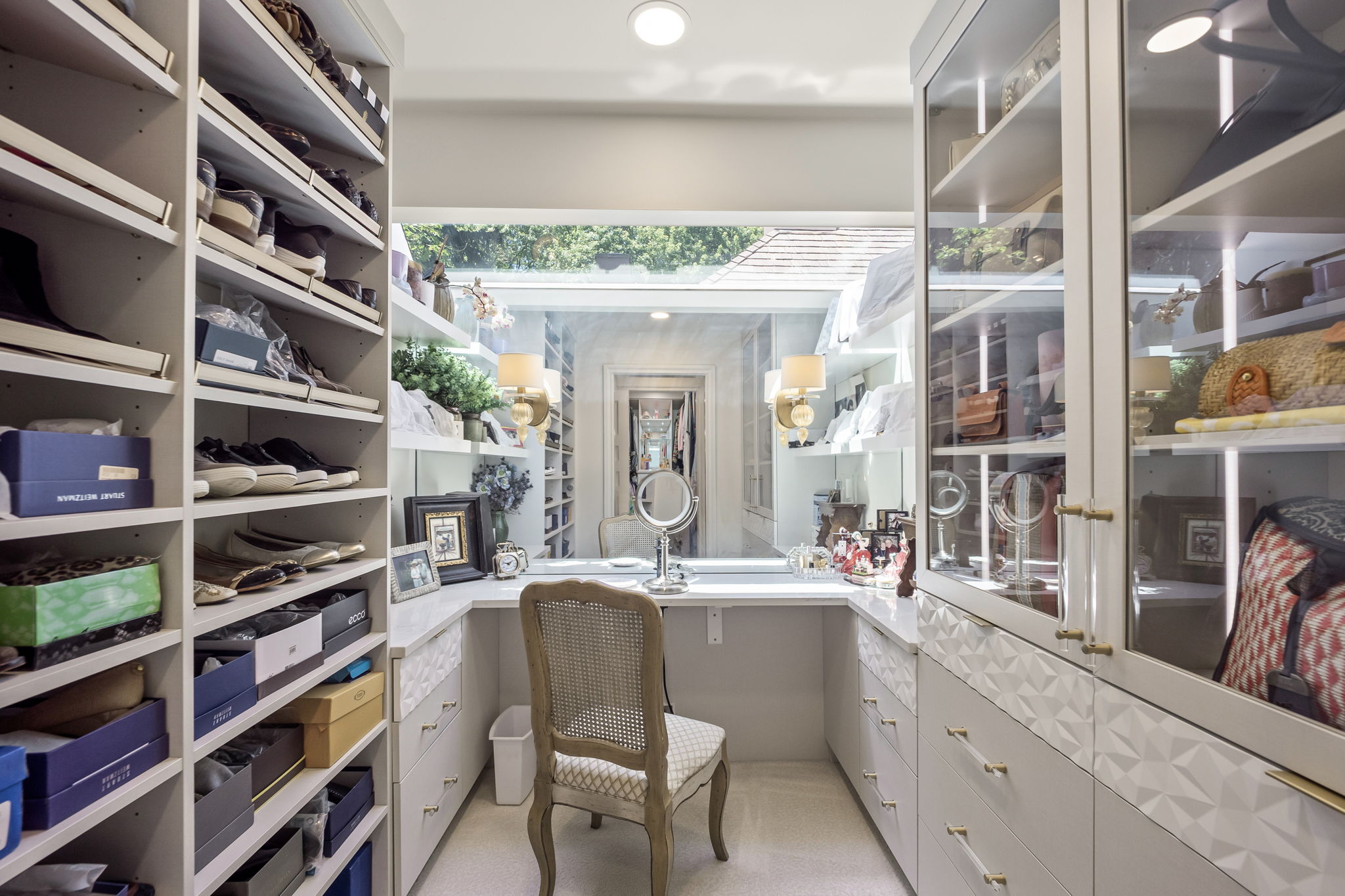 This is a well-organized walk-in closet featuring custom shelving and cabinetry. The closet includes dedicated shoe storage, drawers with decorative fronts, and glass-fronted cabinets for displaying accessories. A vanity area with a chair and mirror is positioned in front of a window, providing natural light.