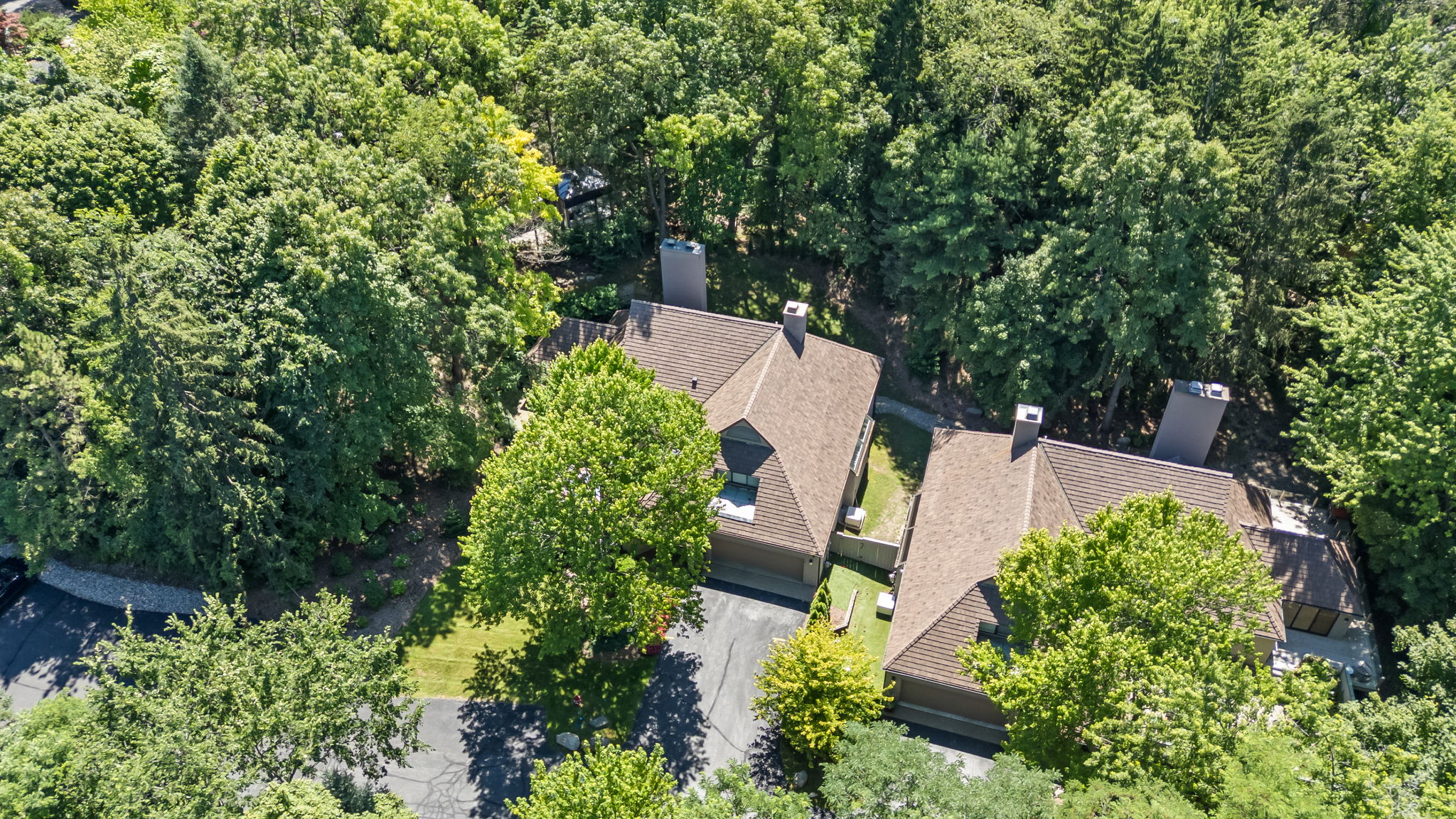 This aerial shot showcases a well-maintained residential property nestled among lush greenery. The house features a brown roof, multiple chimneys, and attached garages. The surrounding trees provide privacy and a sense of seclusion, enhancing the property's appeal.
