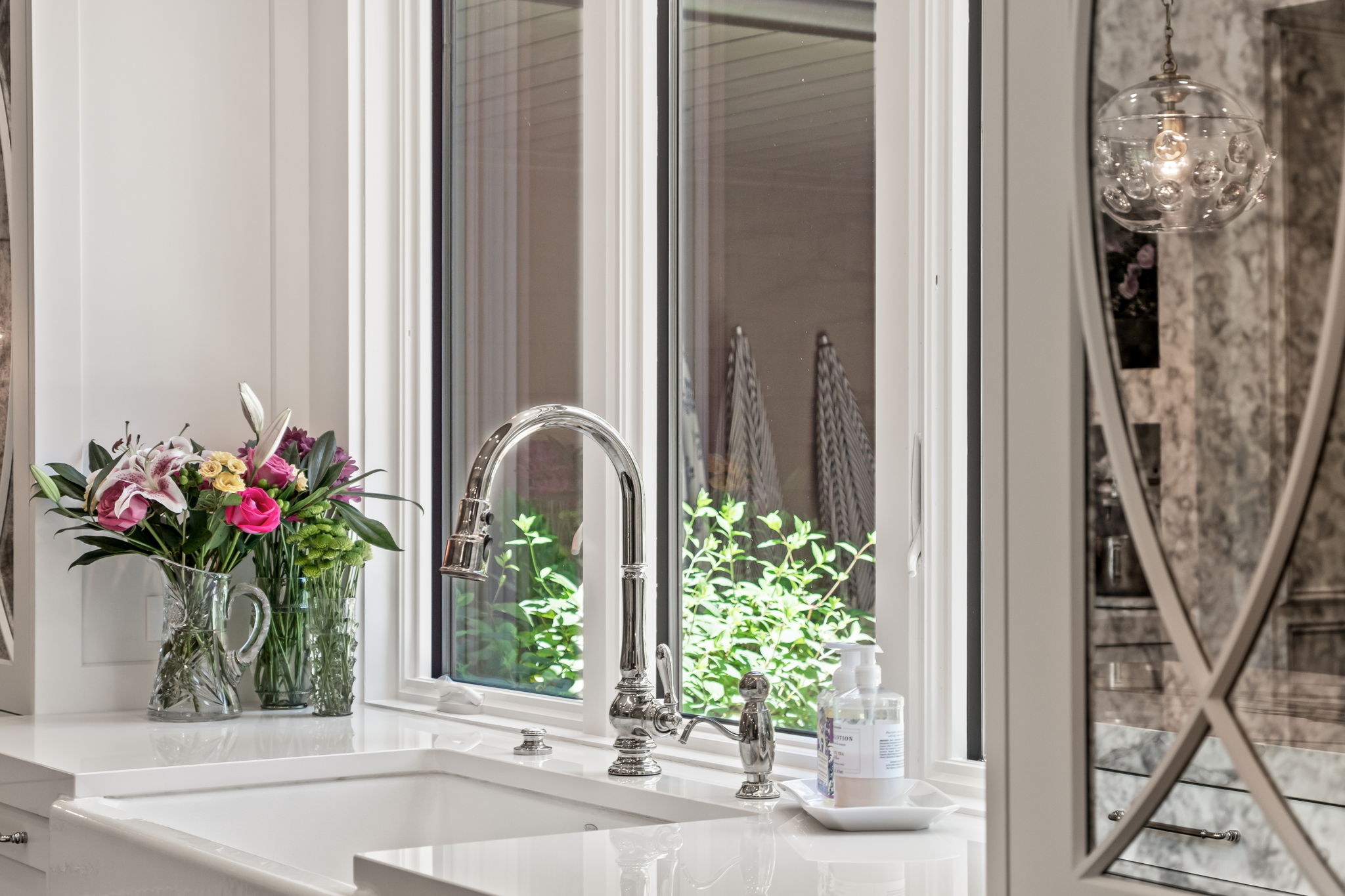 This is a bright and clean kitchen featuring a white farmhouse sink with a chrome faucet and soap dispenser. A vase of colorful flowers sits on the countertop next to the sink, adding a touch of elegance. The window above the sink provides natural light and a view of the greenery outside, enhancing the inviting atmosphere.