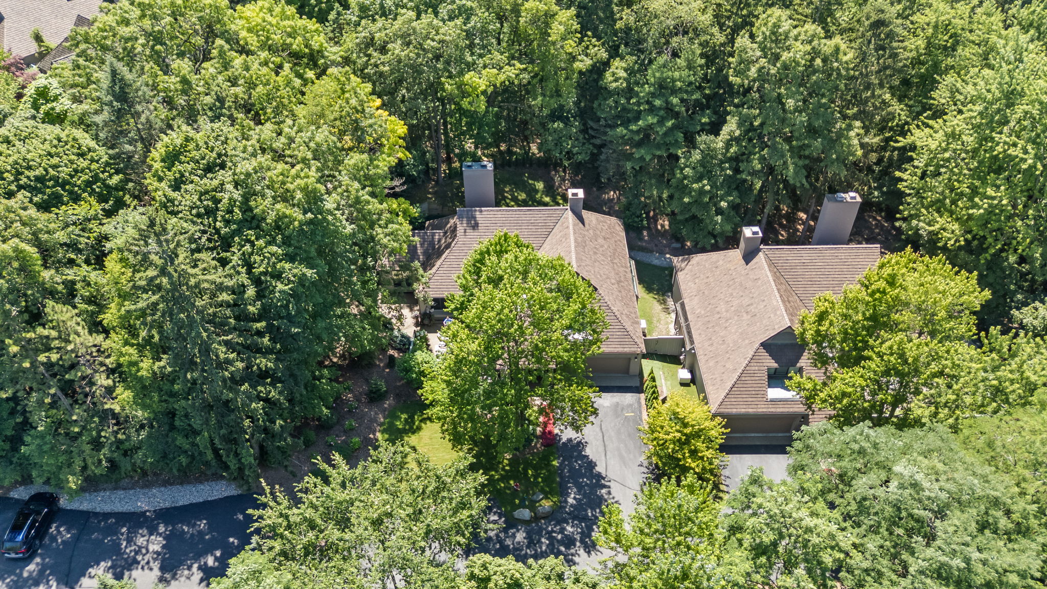 This aerial view showcases a luxurious home nestled among lush greenery. The property features a brown roof, a well-maintained driveway, and mature trees providing ample privacy. The overall impression is one of seclusion and high-end living in a natural setting, with a car parked on the driveway.