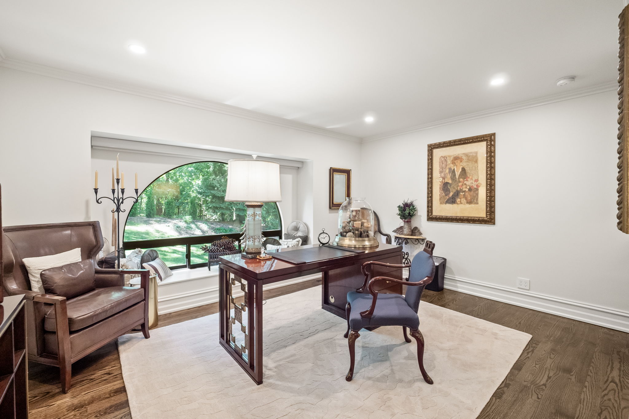 This is an interior shot of a well-appointed home office. The room features a dark wood desk with a blue chair, a leather armchair, and a large arched window providing natural light. The walls are painted white, and the floors are dark hardwood, covered by a light-colored area rug, creating a sophisticated and comfortable workspace.
