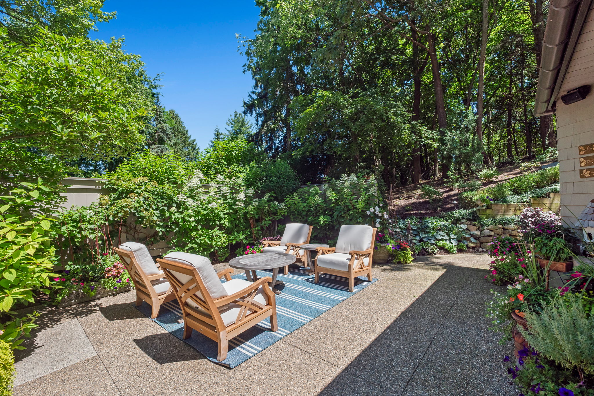 This is a lovely outdoor patio area, perfect for relaxation and entertaining. The space features a set of wooden chairs arranged around a round table, all placed on a blue and white striped rug. The patio is surrounded by lush greenery, including trees, bushes, and potted plants, creating a private and serene atmosphere.