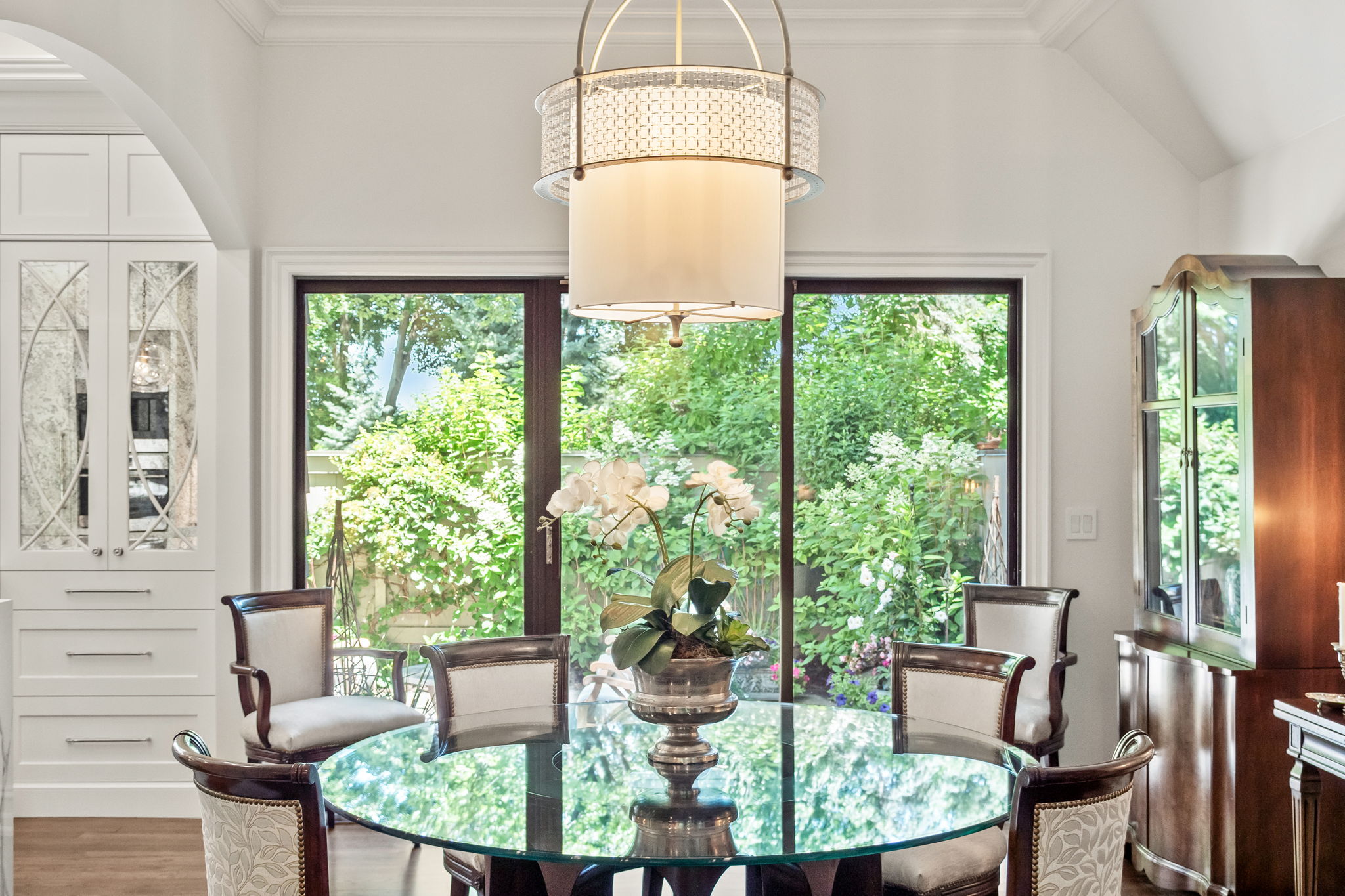 This is an interior shot of a dining room featuring a round glass table surrounded by upholstered chairs with dark wood frames. A modern chandelier hangs above the table, and large windows provide a view of a lush green garden. Built-in cabinetry and a dark wood hutch add to the room's elegance and functionality.
