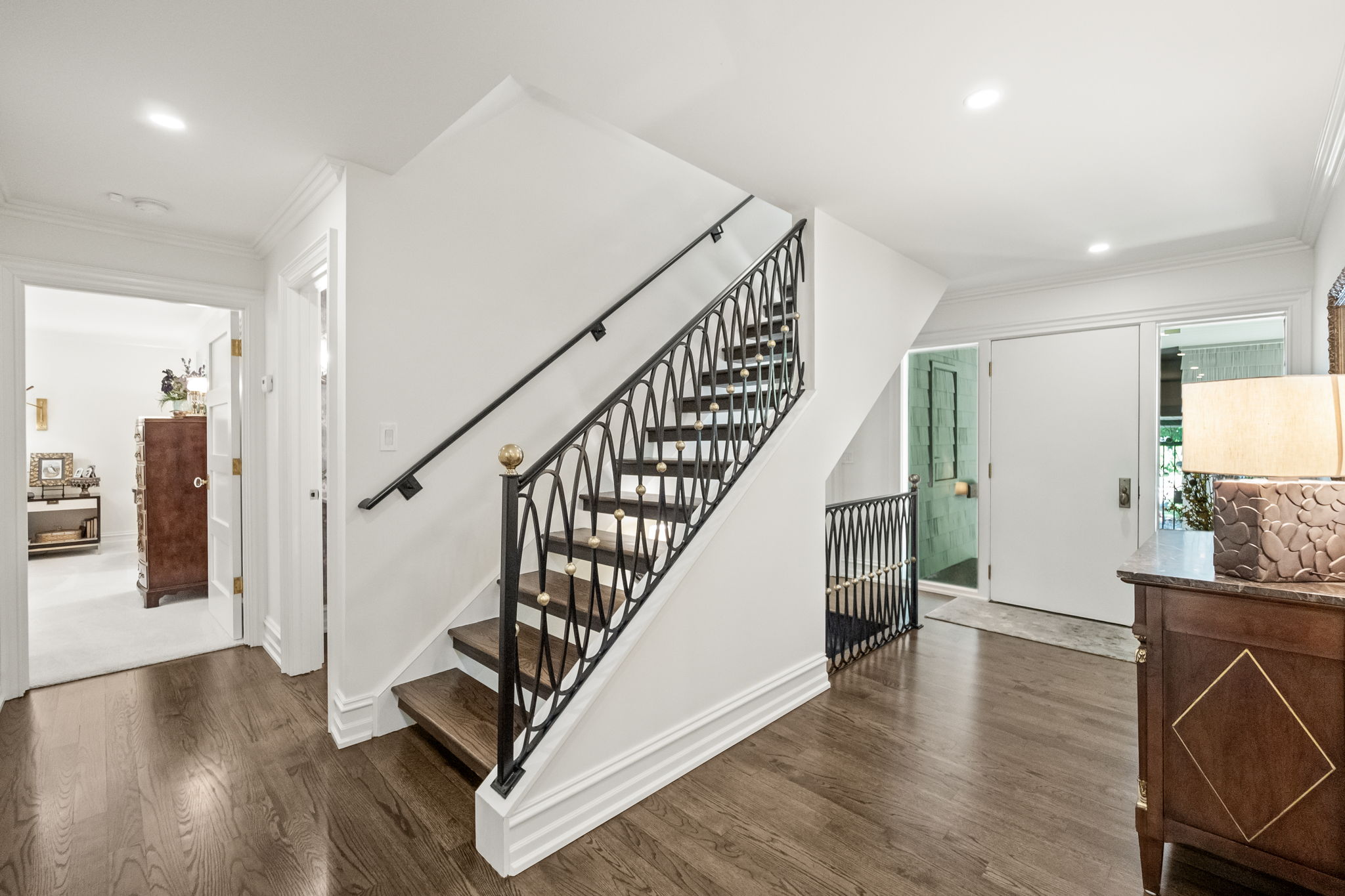 This interior shot showcases a well-lit hallway with elegant wooden flooring and a staircase featuring a decorative black iron railing with gold accents. The walls are painted in a neutral tone, creating a bright and inviting space. The hallway leads to various rooms, suggesting a spacious and well-designed home.