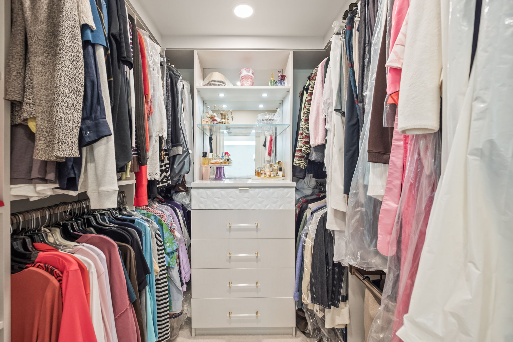 This is a well-organized walk-in closet featuring clothing racks filled with garments on both sides and a central built-in dresser with drawers and shelving. The closet is brightly lit, creating a clean and luxurious feel. The perspective is from the entrance of the closet, showcasing the depth and storage capacity.